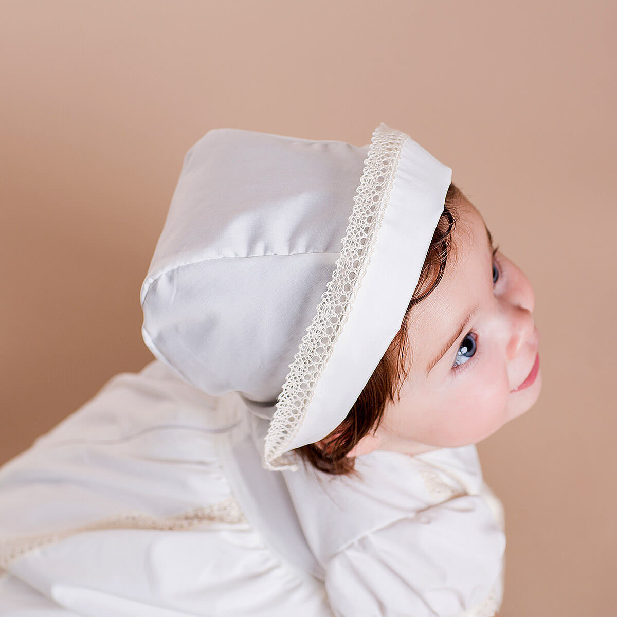Child wearing a Baptism Bonnett with lace trim against a beige background