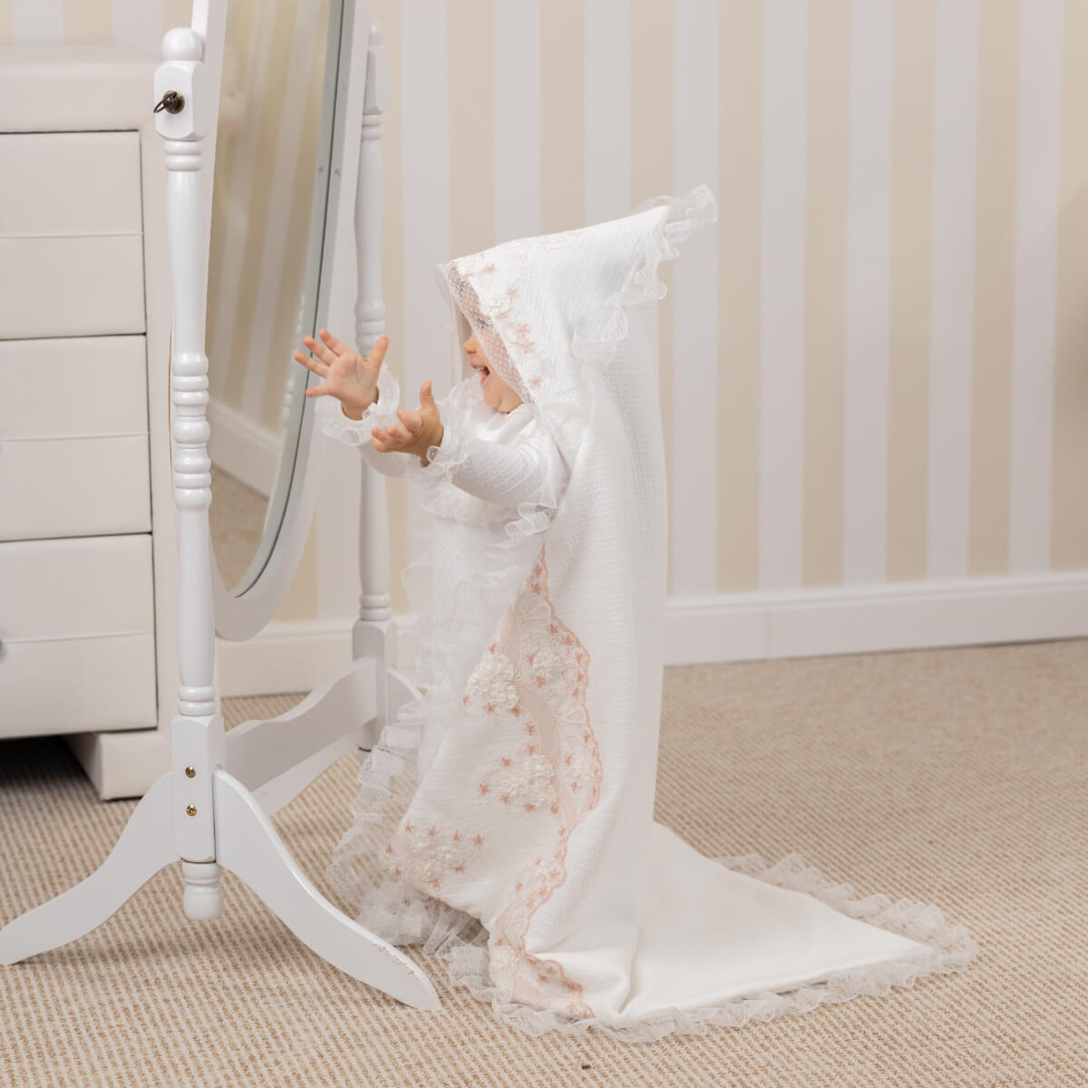 Baby in baptism blanket with a long train standing next to a white stand.