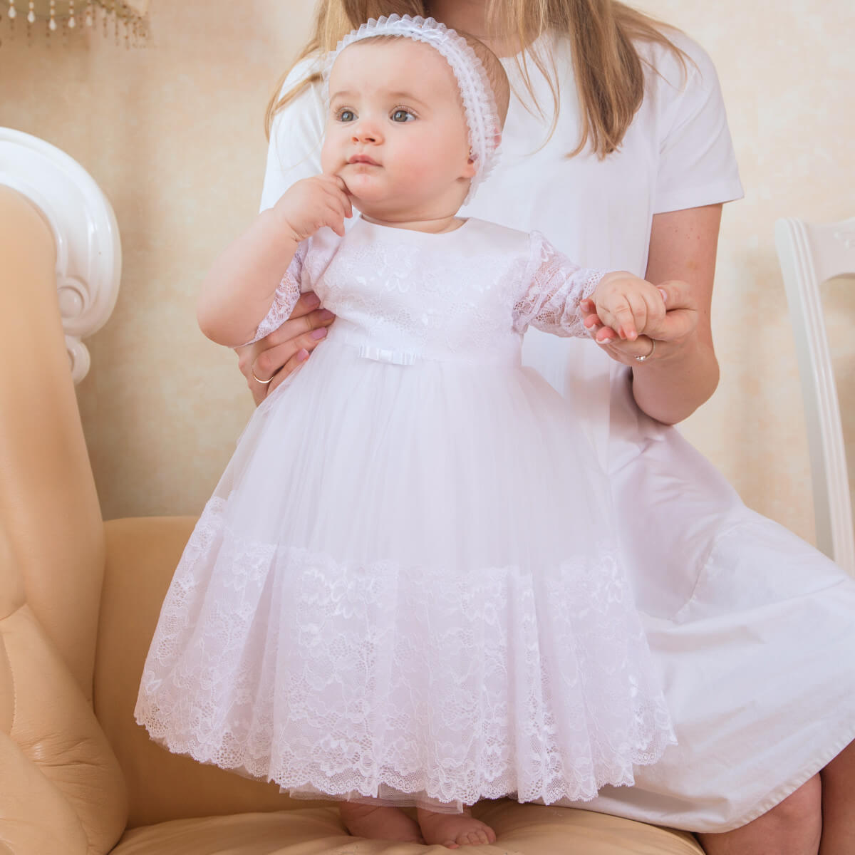 Baby in a Baptism Tulle Lace Dress being held by an adult, sitting on a beige chair.
