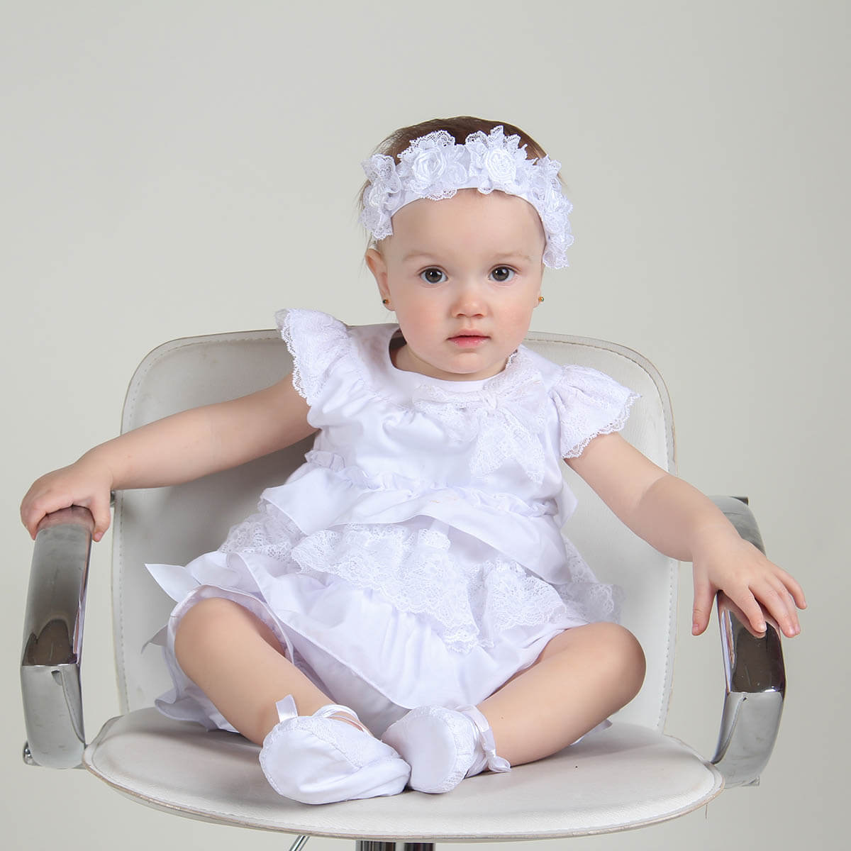Baby in a Christening Lace Dress and headband sitting on a chair against a plain background