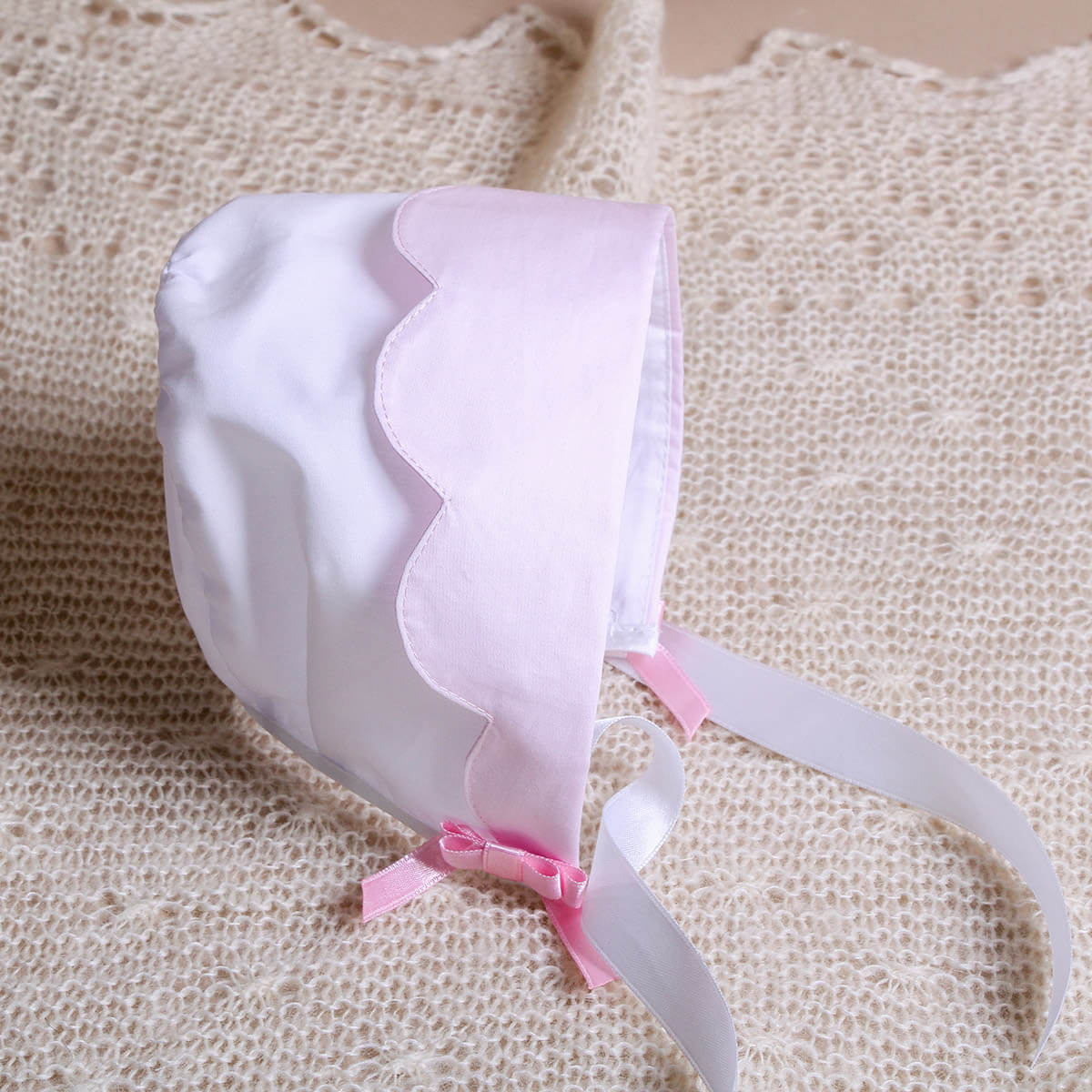 White Cotton Baptism Bonnet with pink trim on a textured beige background