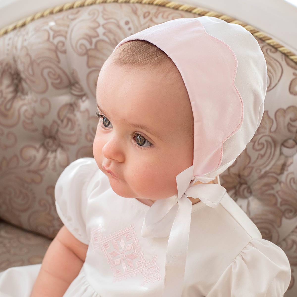 Baby wearing a p Cotton Baptism Bonnet sitting on a patterned chair.