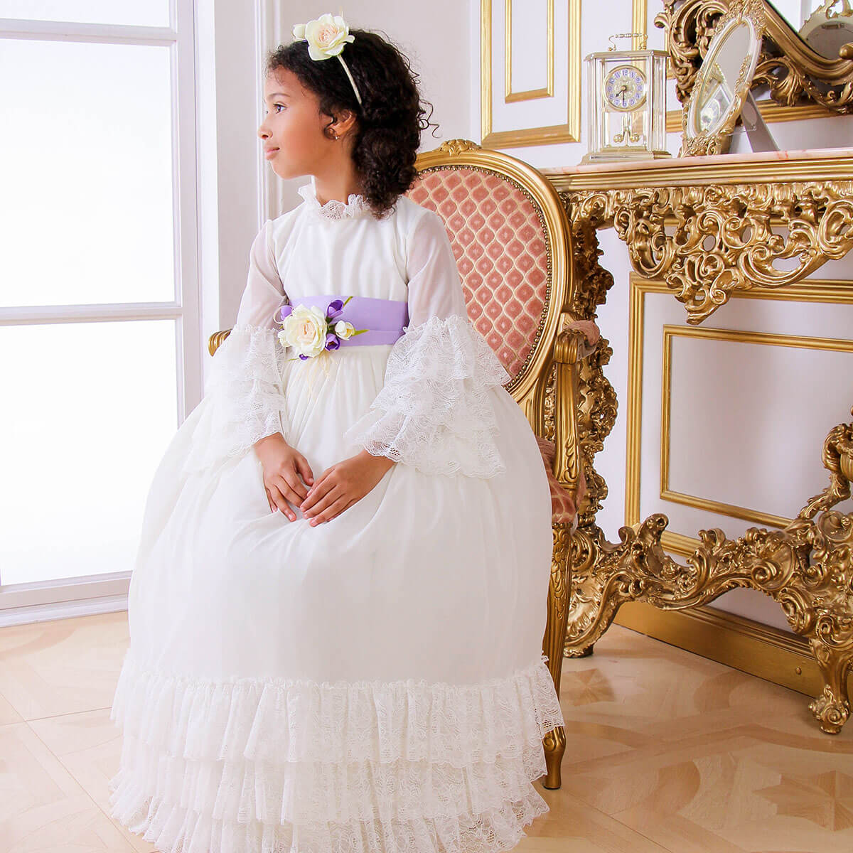 Young girl in Boho First Communion Dress with floral accents sitting in an ornate chair.
