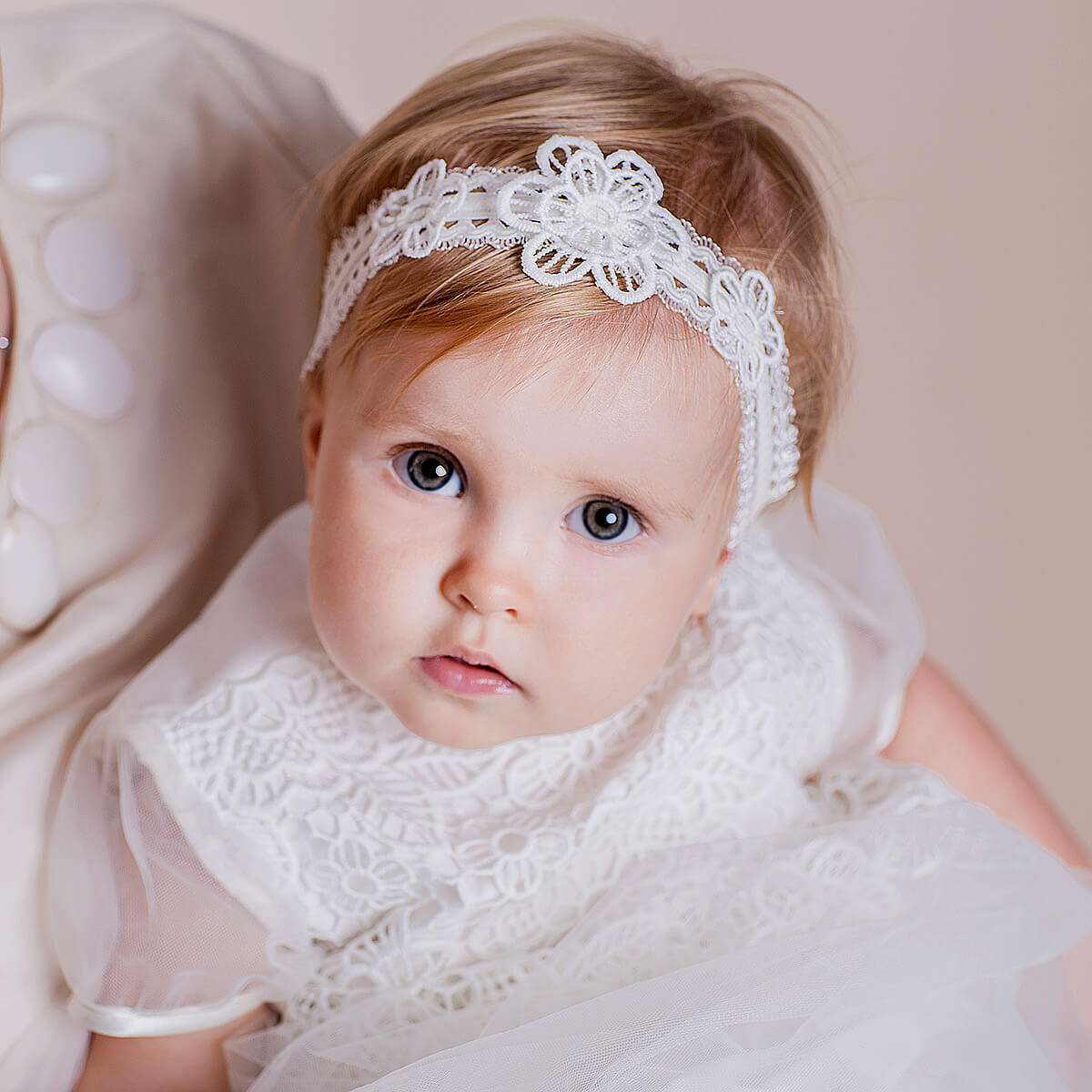 Baby wearing a white lace headband and dress against a neutral background