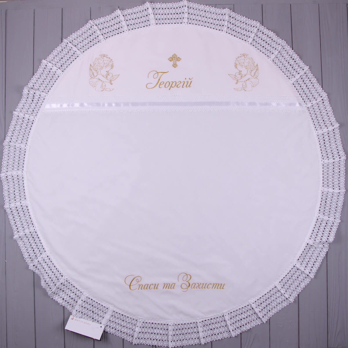 Round white embroidered christening blanket with decorative border on a wooden surface