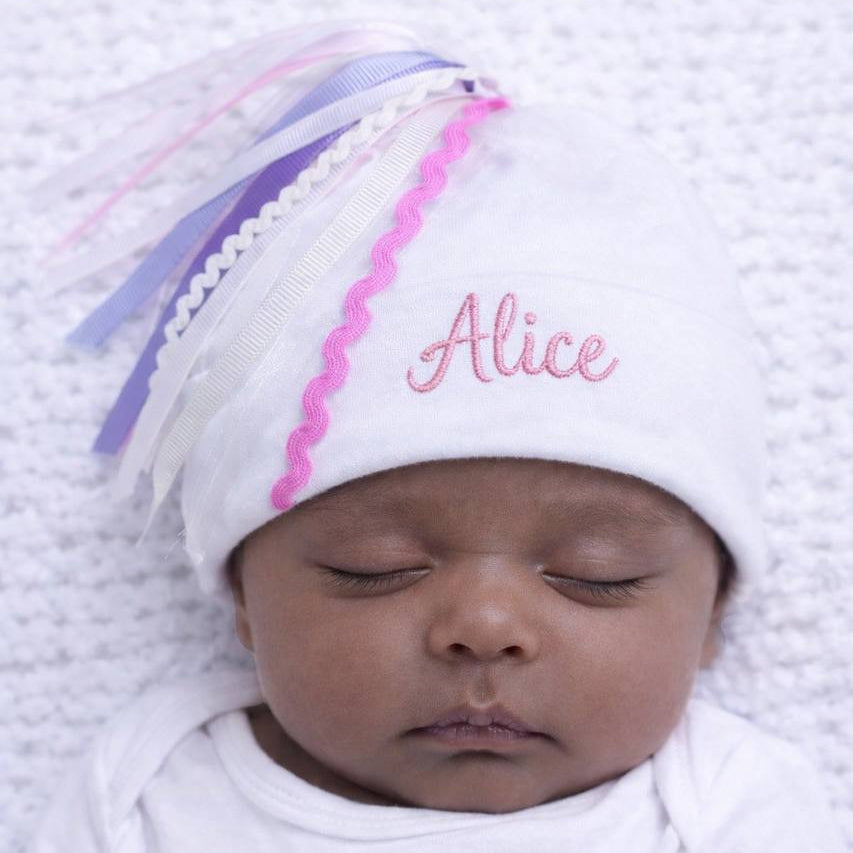 Newborn baby wearing a white hat with colorful tassels and the name 'Alice' embroidered on it, lying on a white blanket.
