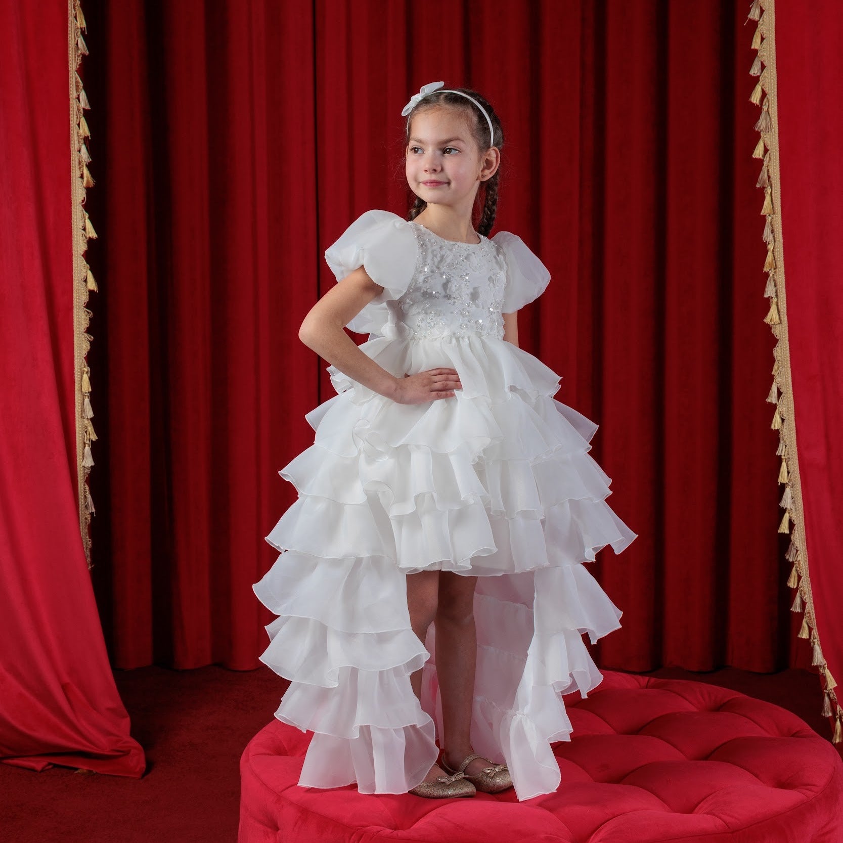 Young girl in a white dress standing on a red ottoman with red curtains in the background