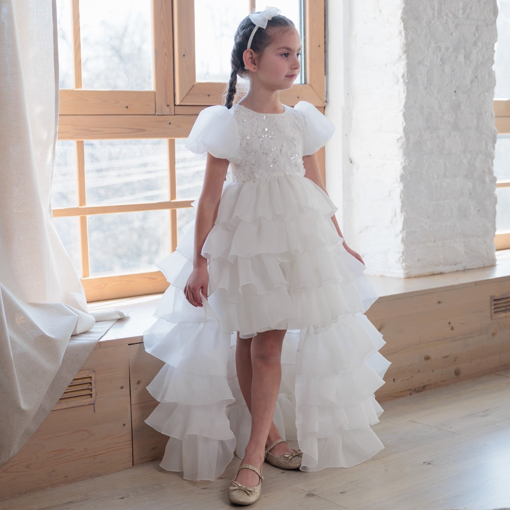 Young girl in a white dress standing in a bright room with large windows.