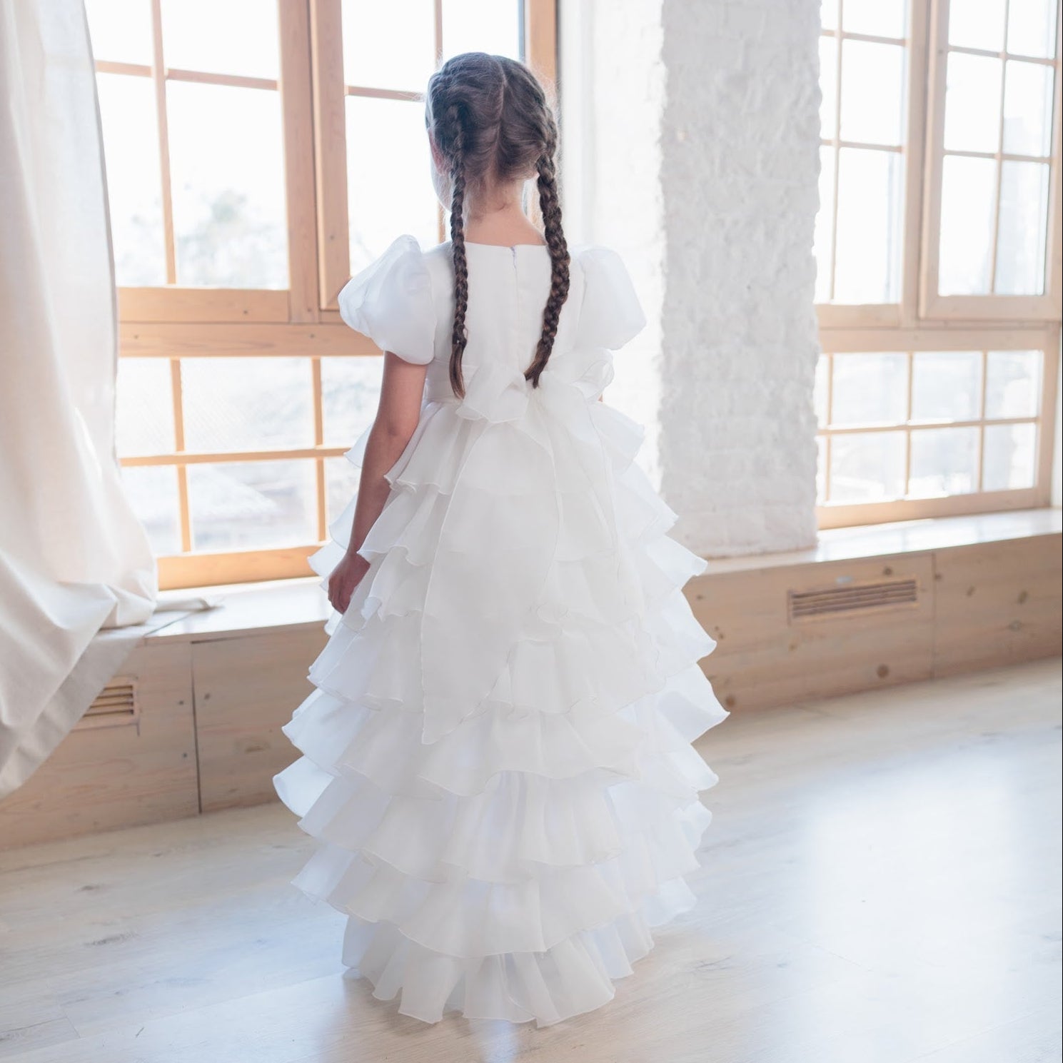 Young girl in a white dress standing in a bright room with large windows.