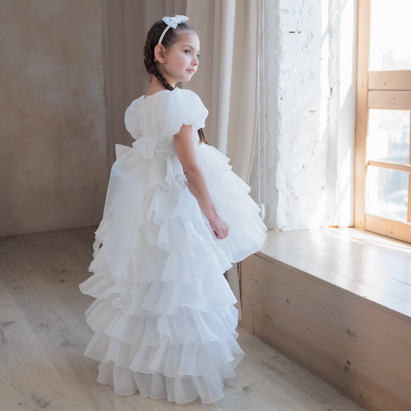 Young girl in a white dress standing in a softly lit room with large windows.