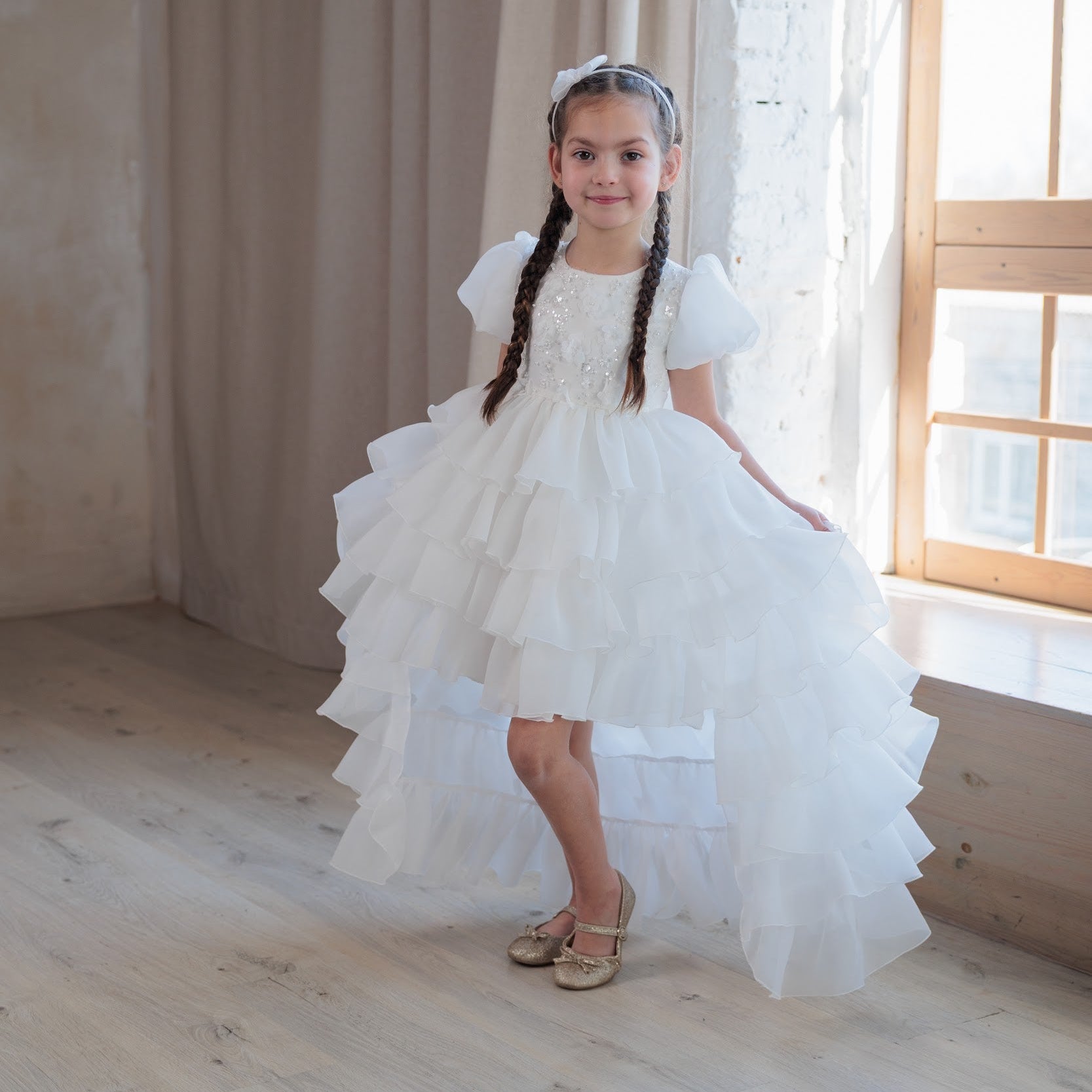 Young girl in a white dress standing in a softly lit room with curtains and a window.