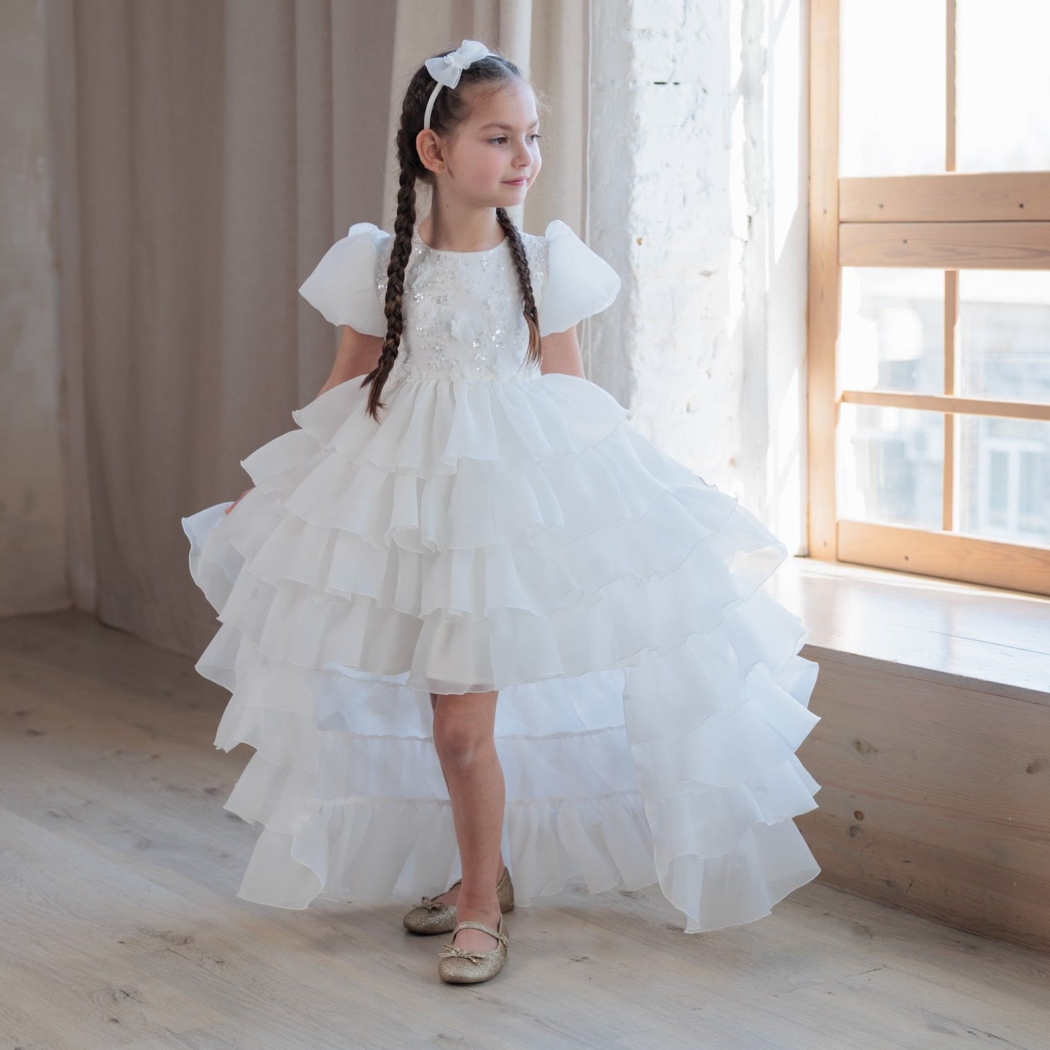 Young girl in a white dress standing in a bright room with large windows.