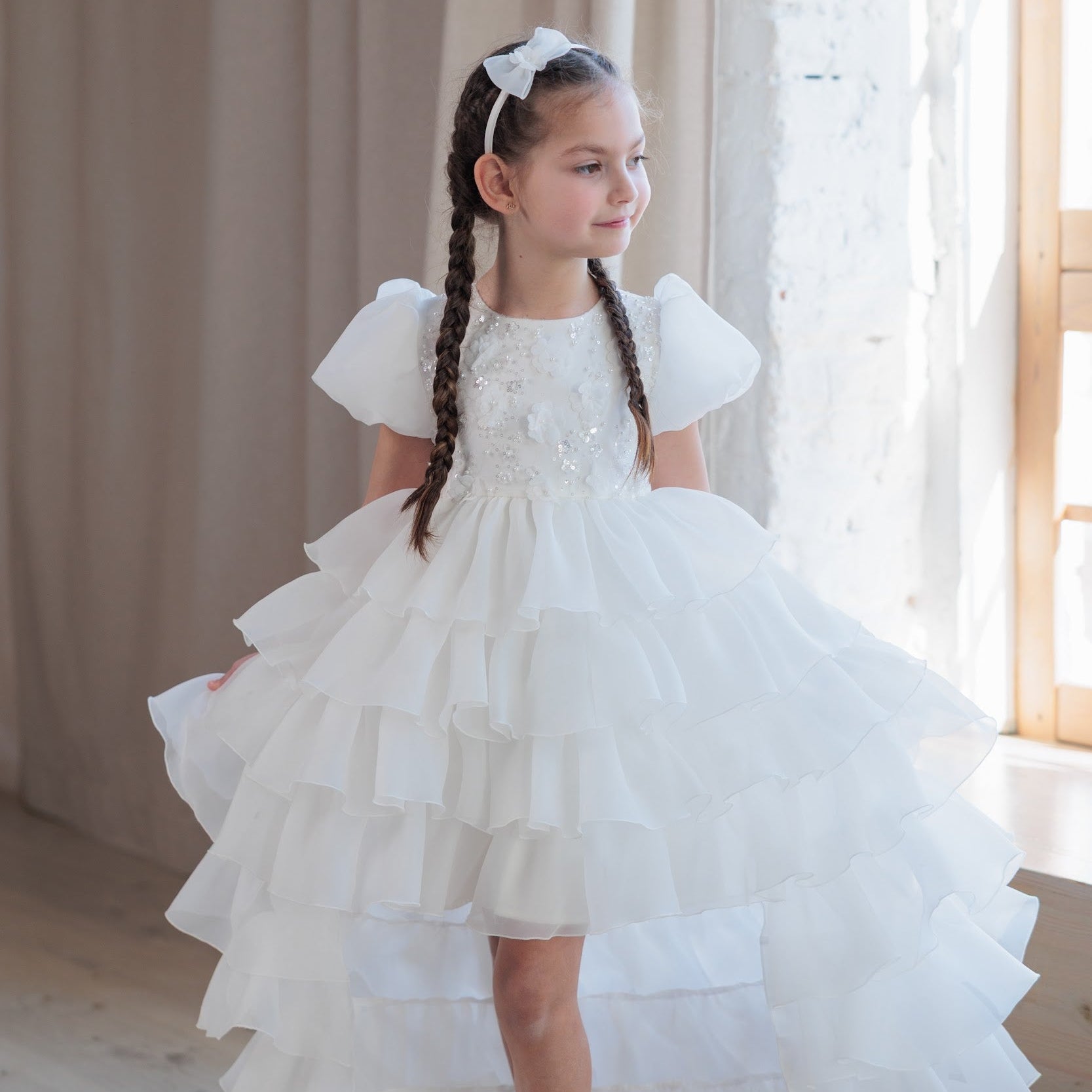 Young girl in a white dress standing in a softly lit room with curtains.