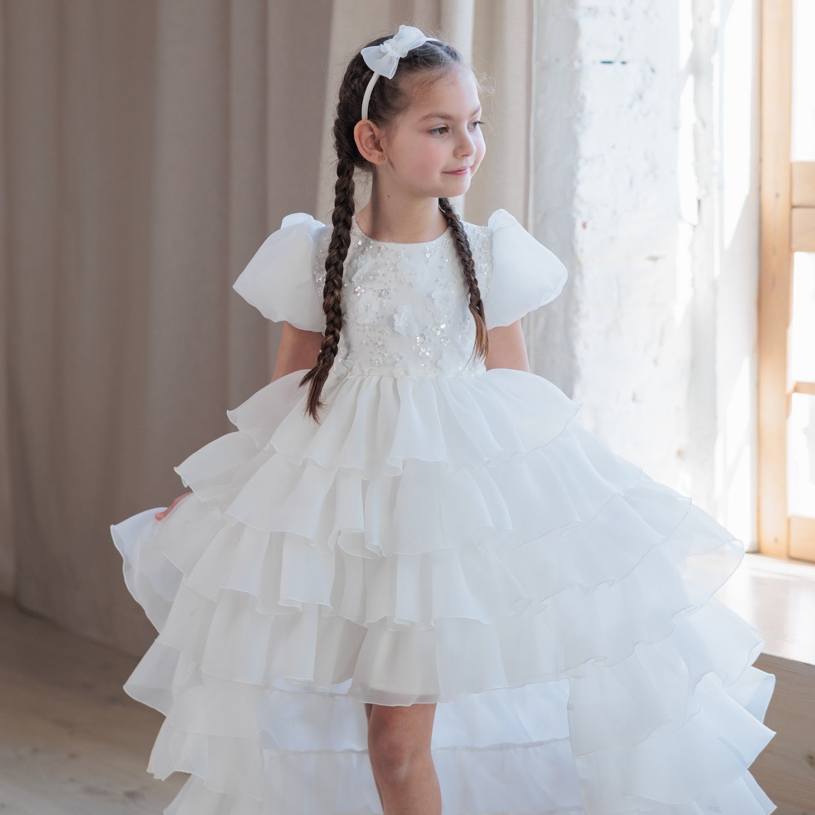 Young girl in a white dress standing in a softly lit room with curtains.