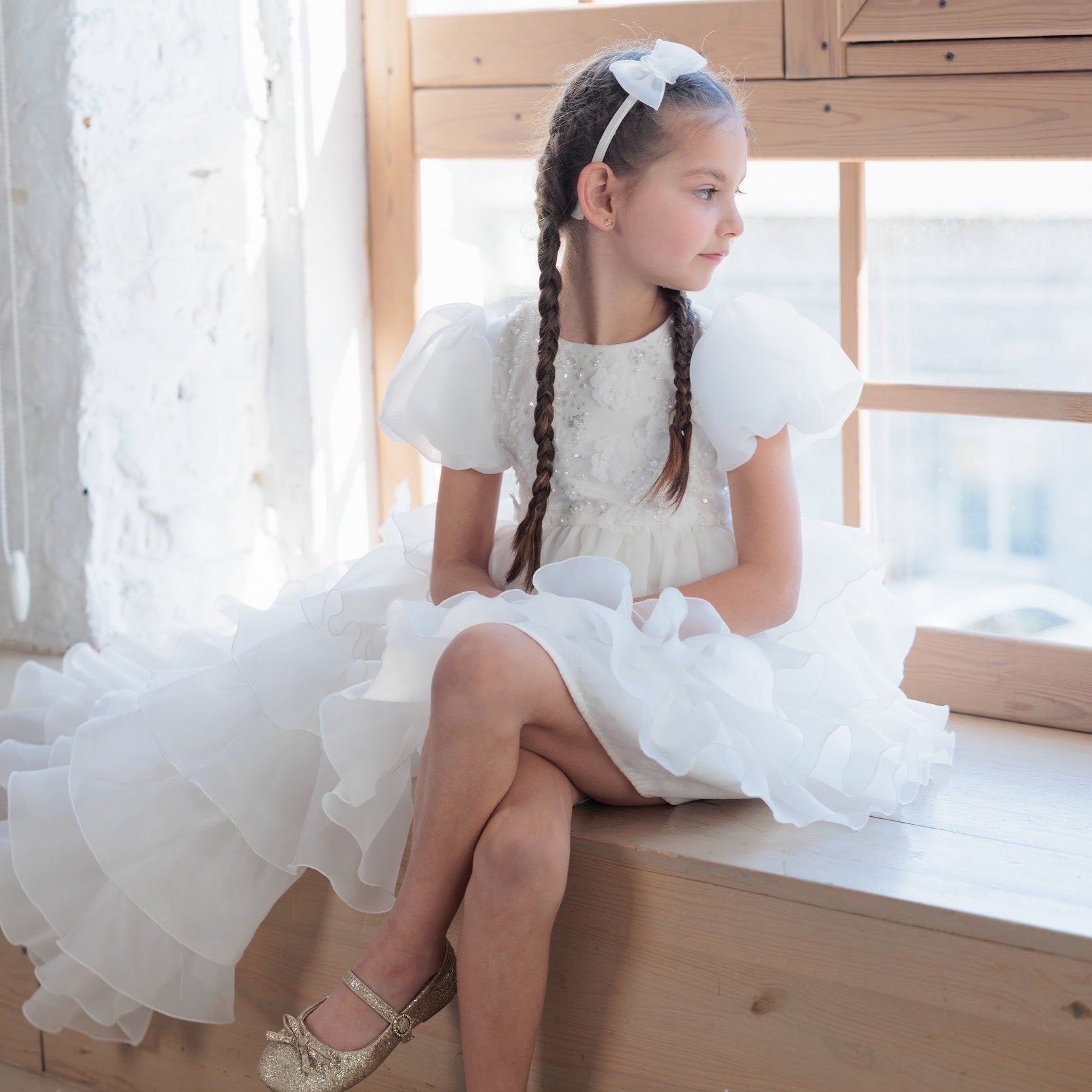 Young girl in a white dress sitting by a window with a wooden frame.