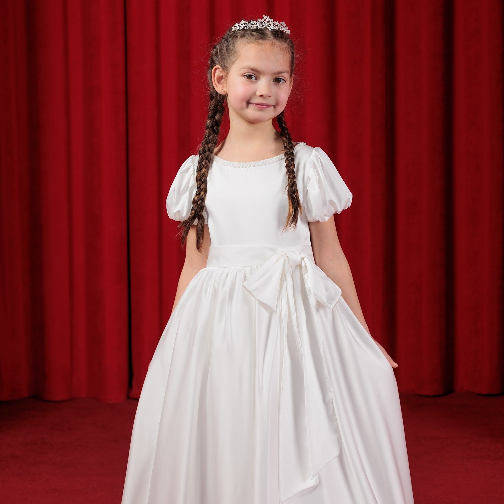 Young girl in a white silk simple flower girl dress with braided hair against a red curtain background