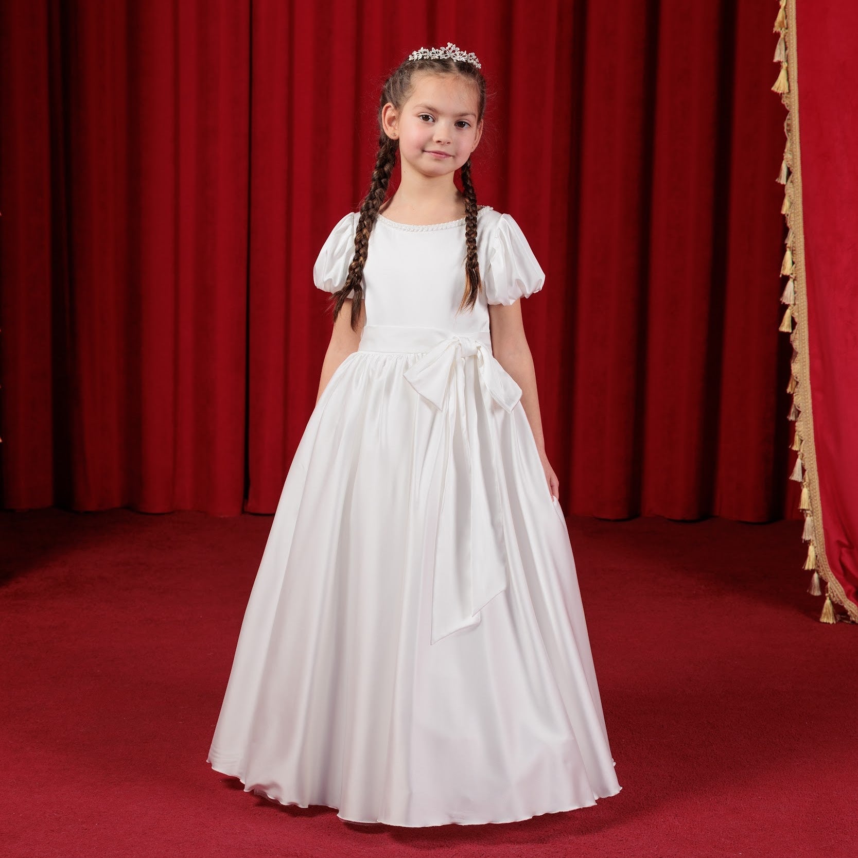 Young girl in a white  silk simple flower girl dress standing in front of red curtains