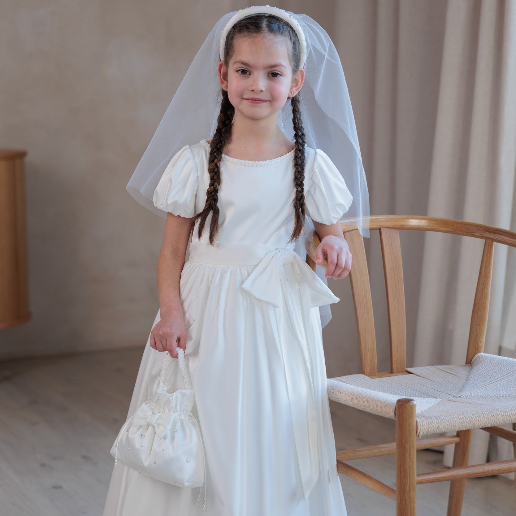 Young girl in a white silk communion dress with a veil standing indoors.
