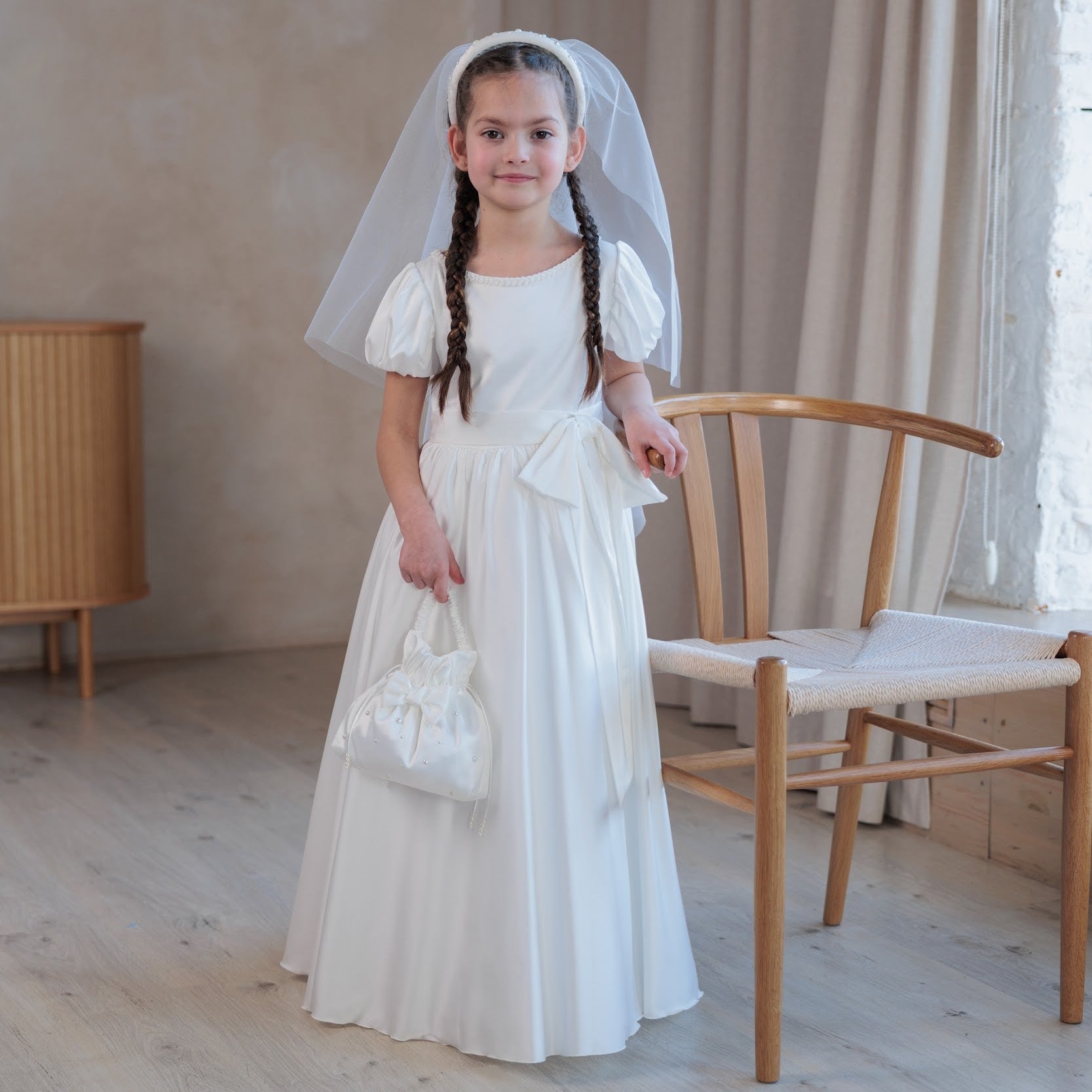 Young girl in a white silk communion dress with a veil standing in a room with wooden floor and furniture.