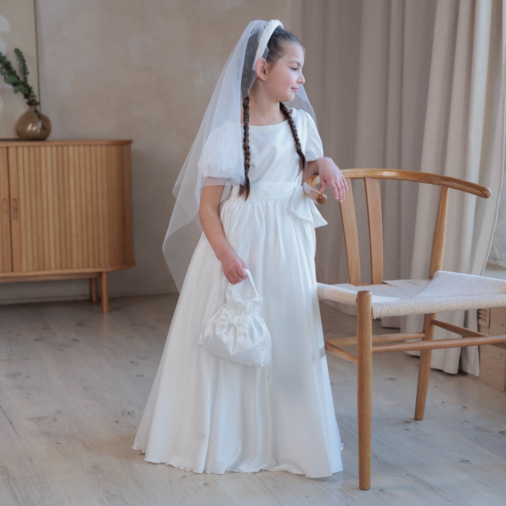 Young girl in a white silk communion dress with a veil standing in a room with wooden furniture.