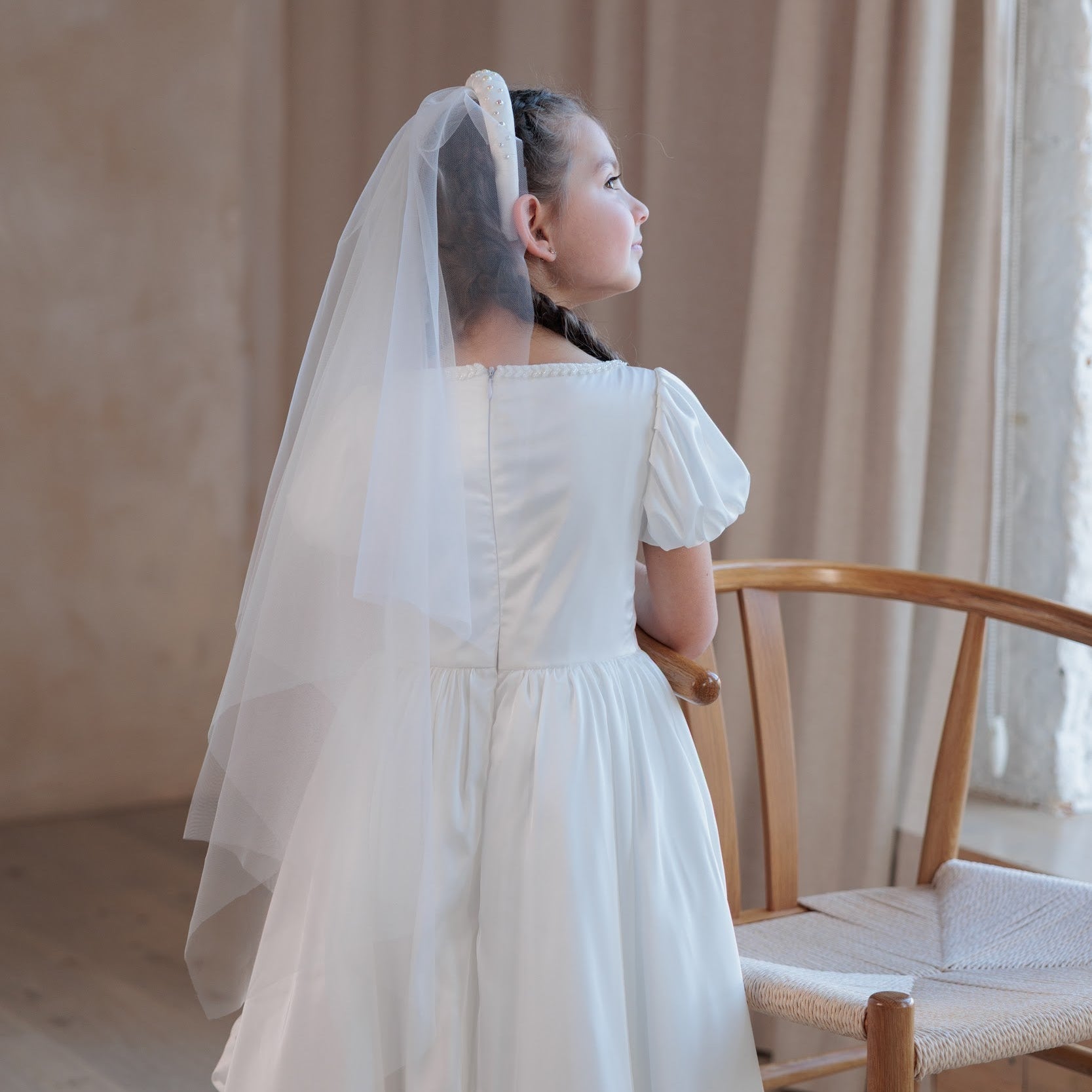Woman in a white silk communion dress with a veil standing in a room with curtains and a chair.