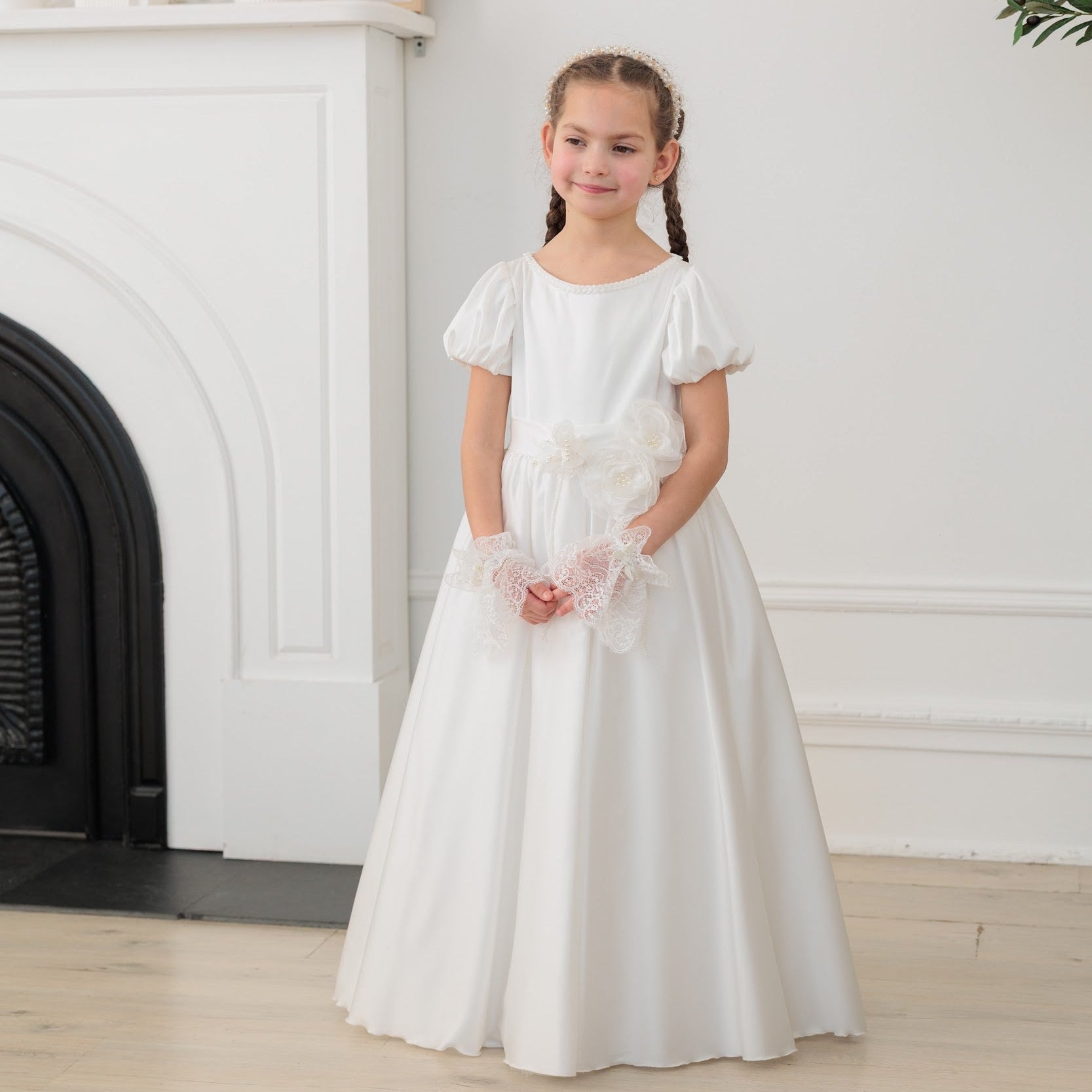 Young girl in a white silk communion dress standing in a room with a fireplace and decorative items.