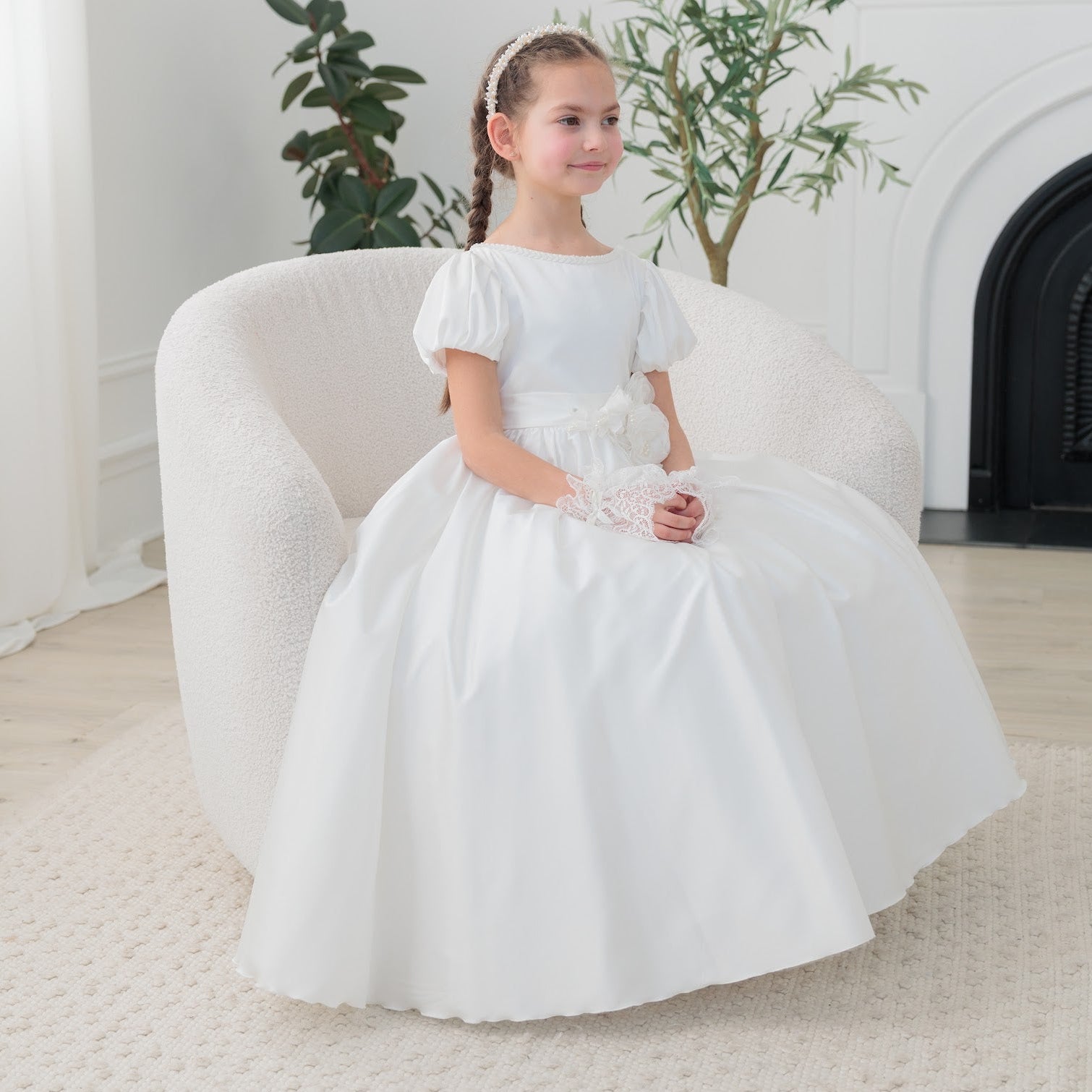 Young girl in a white silk communion dress sitting on a white chair in a room with plants and a fireplace.