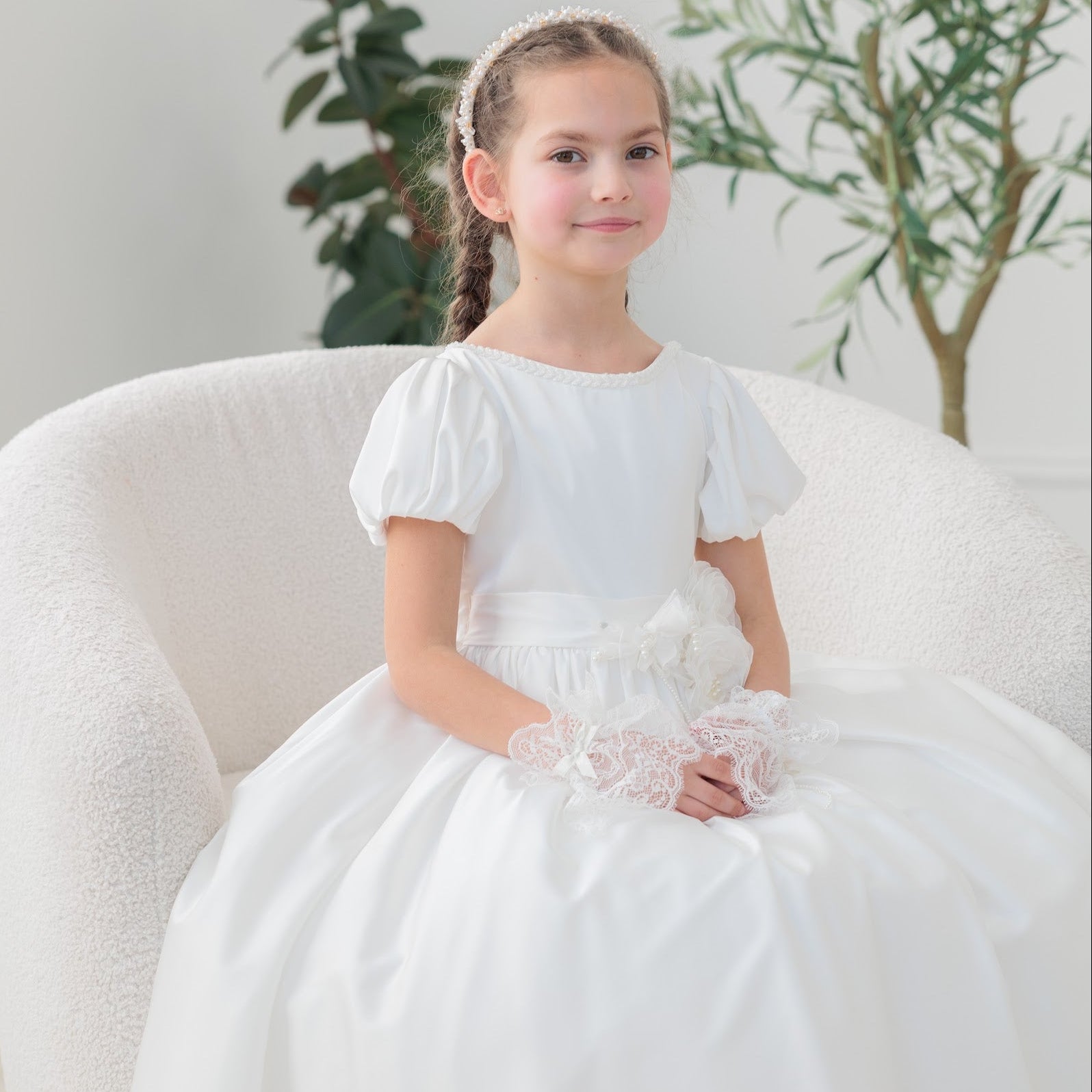 Young girl in a white silk communion dress sitting on a white chair with plants in the background