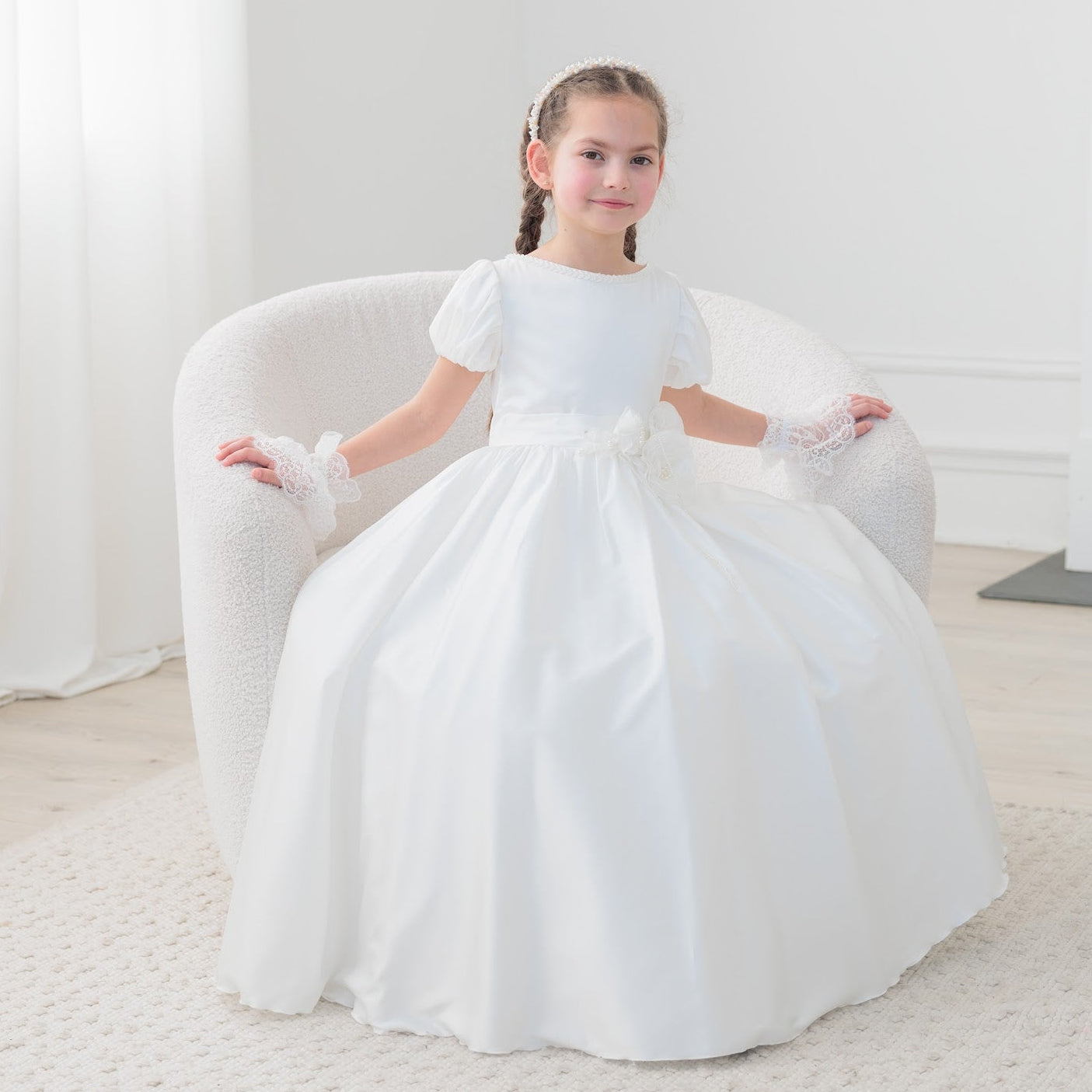 Young girl in a white silk communion dress sitting on a white chair in a bright room.