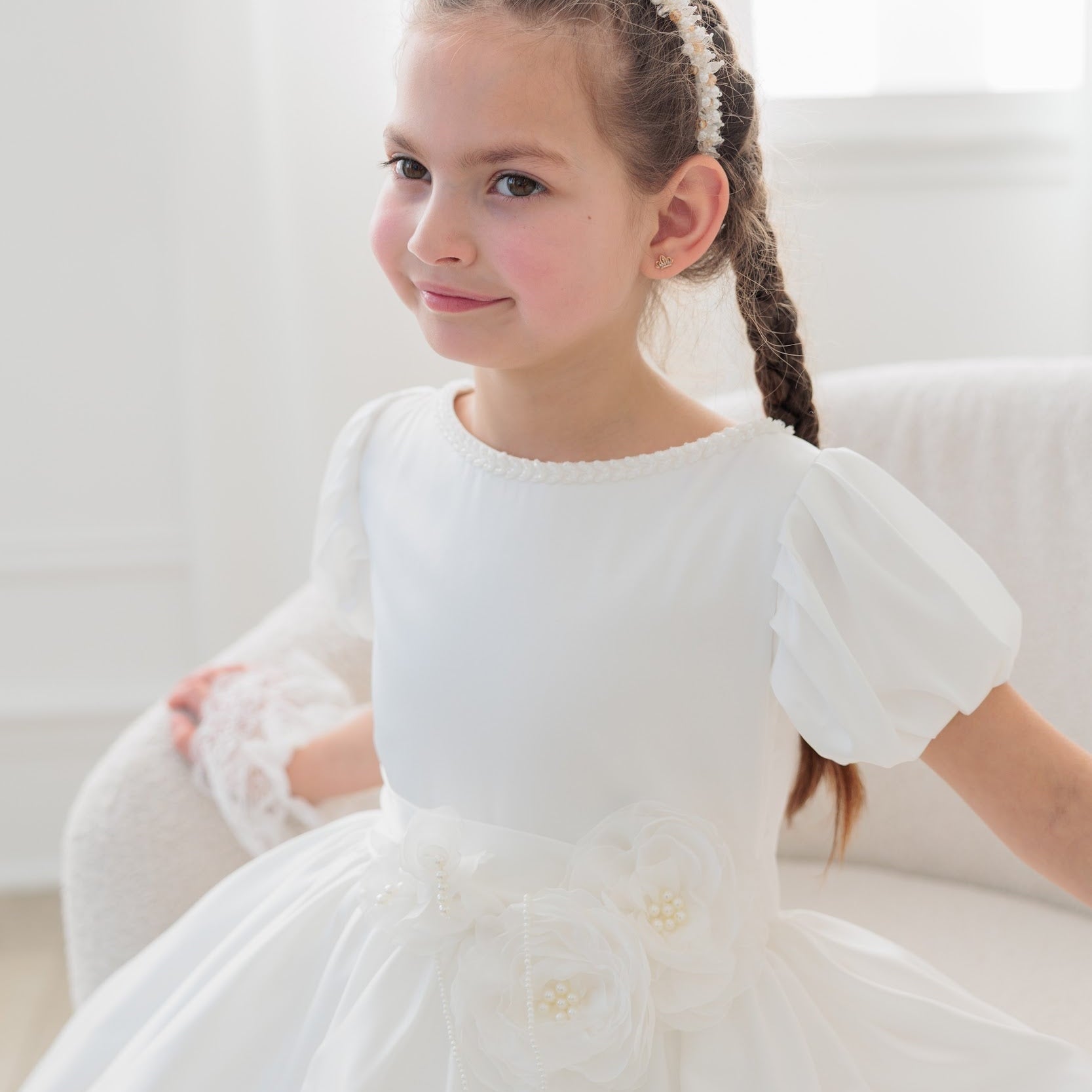 Young girl in a white silk communion dress sitting on a white couch with a white curtain background