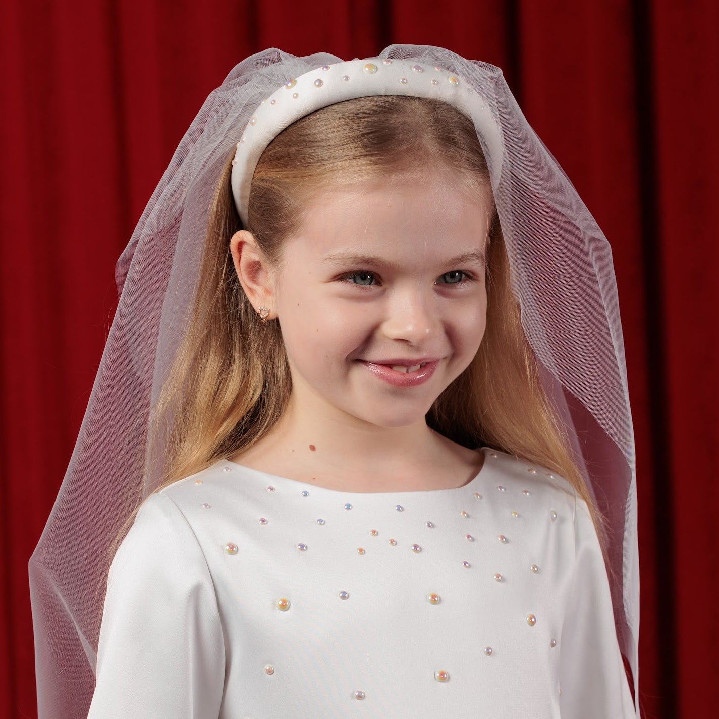 Young girl in a white communion dress with a veil against a red curtain background