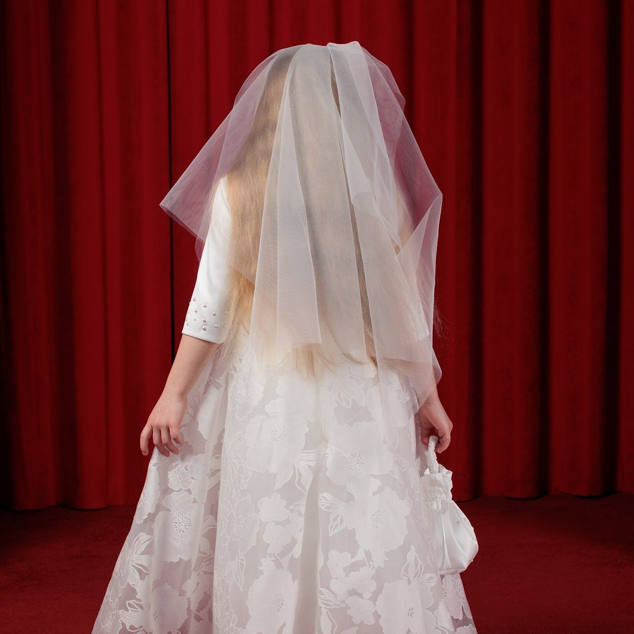 Person wearing a white lace dress and communion veil against a red curtain background