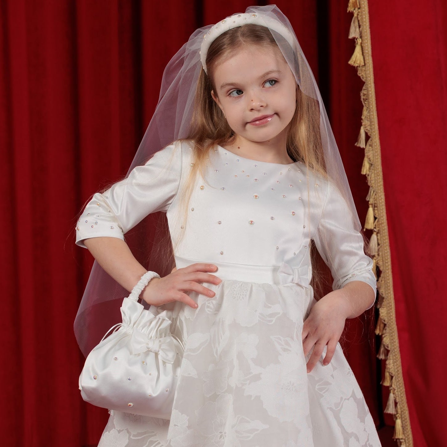 Young girl in a white lace dress with a veil standing in front of red curtains.