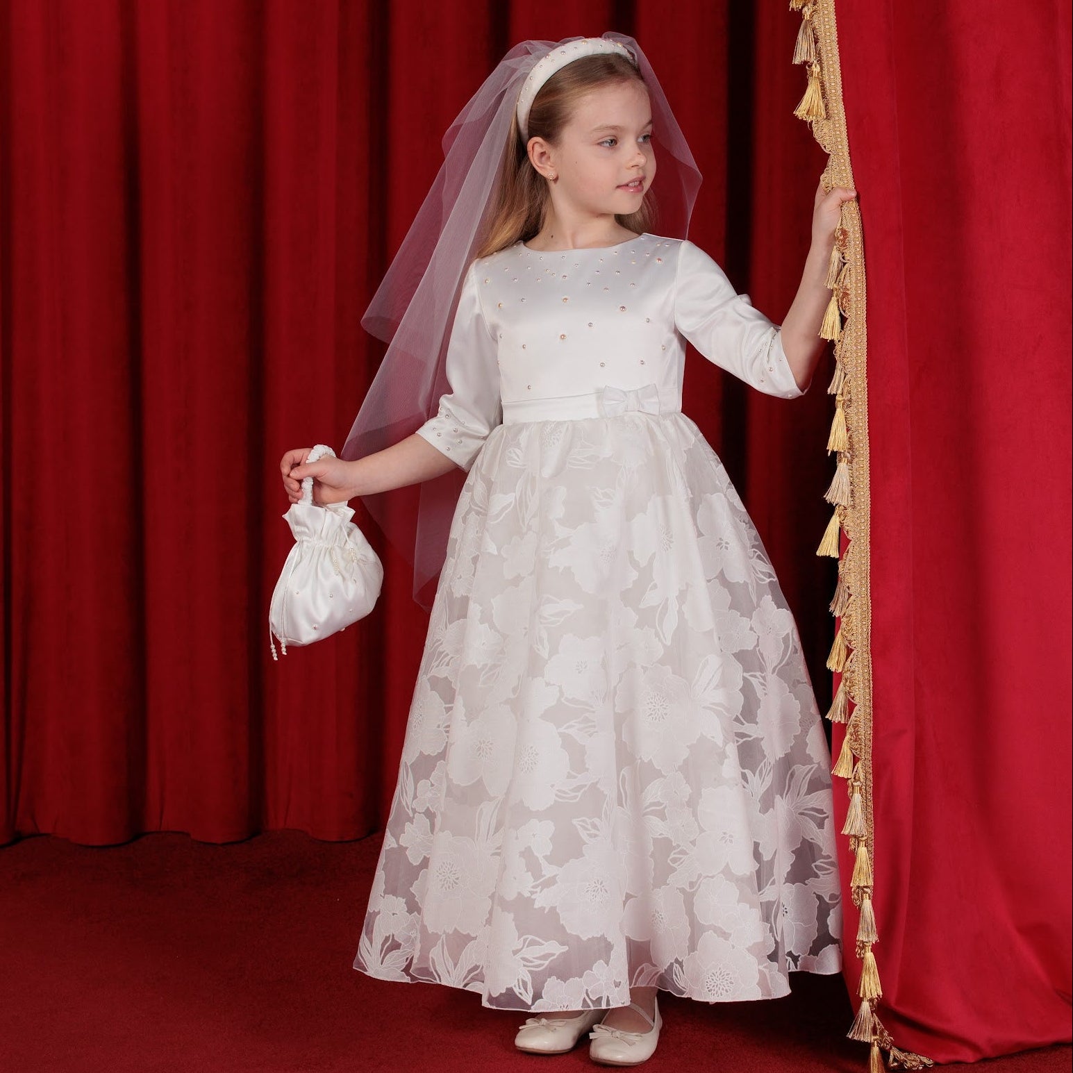 Young girl in a white communion dress with floral pattern standing in front of red curtains.