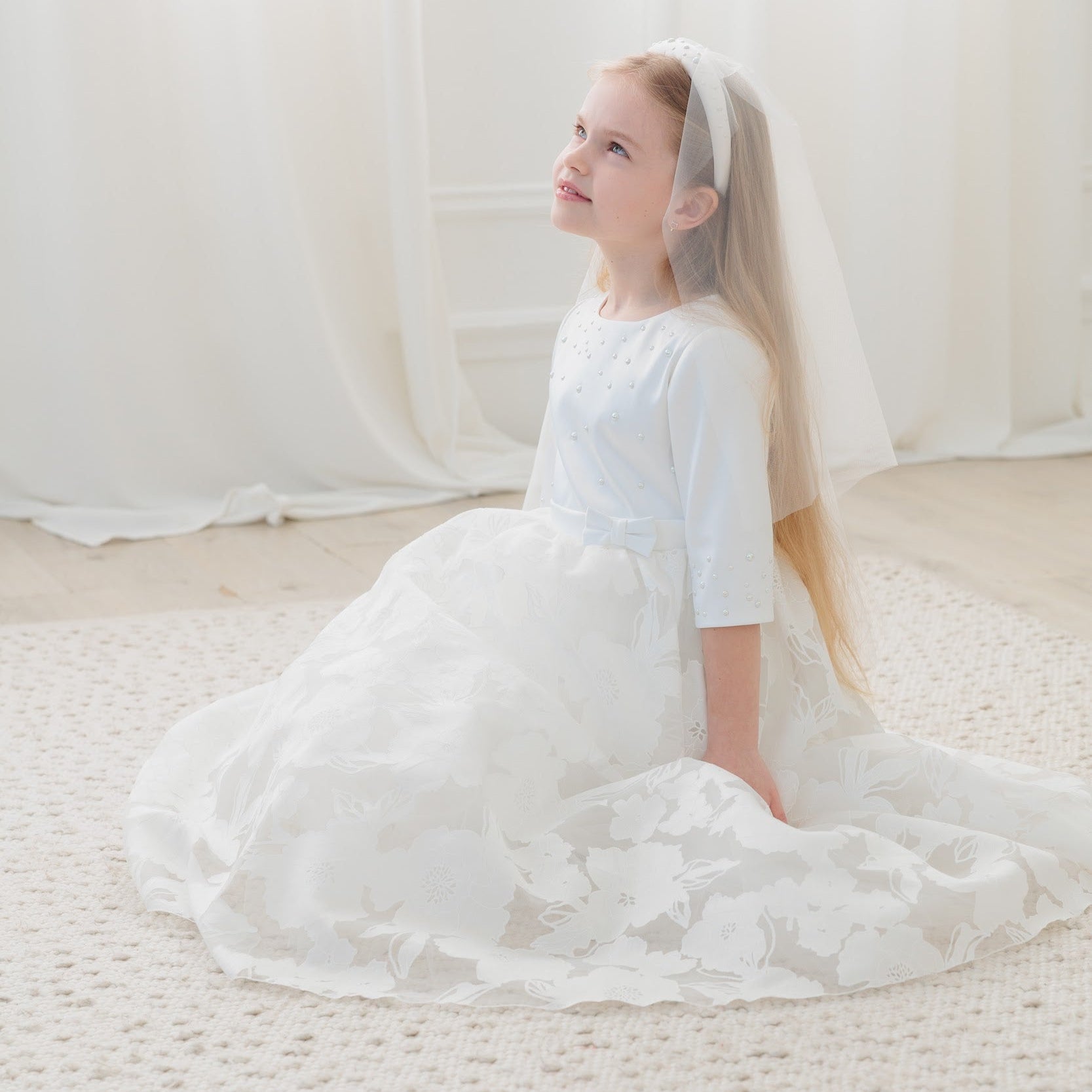 Young girl in a white dress with a veil sitting on a light-colored carpet.