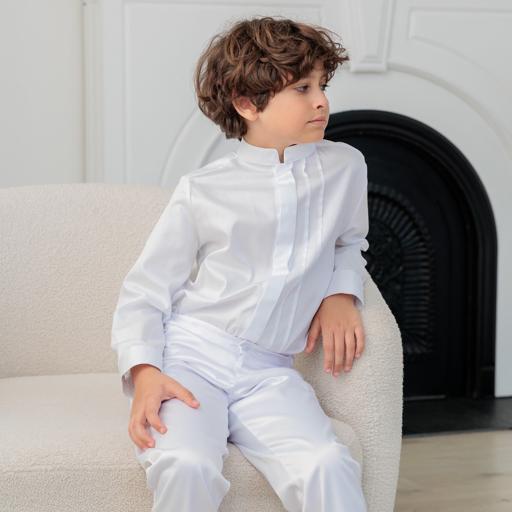 Young boy in a white communion outfit sitting on a couch in a room with a fireplace.