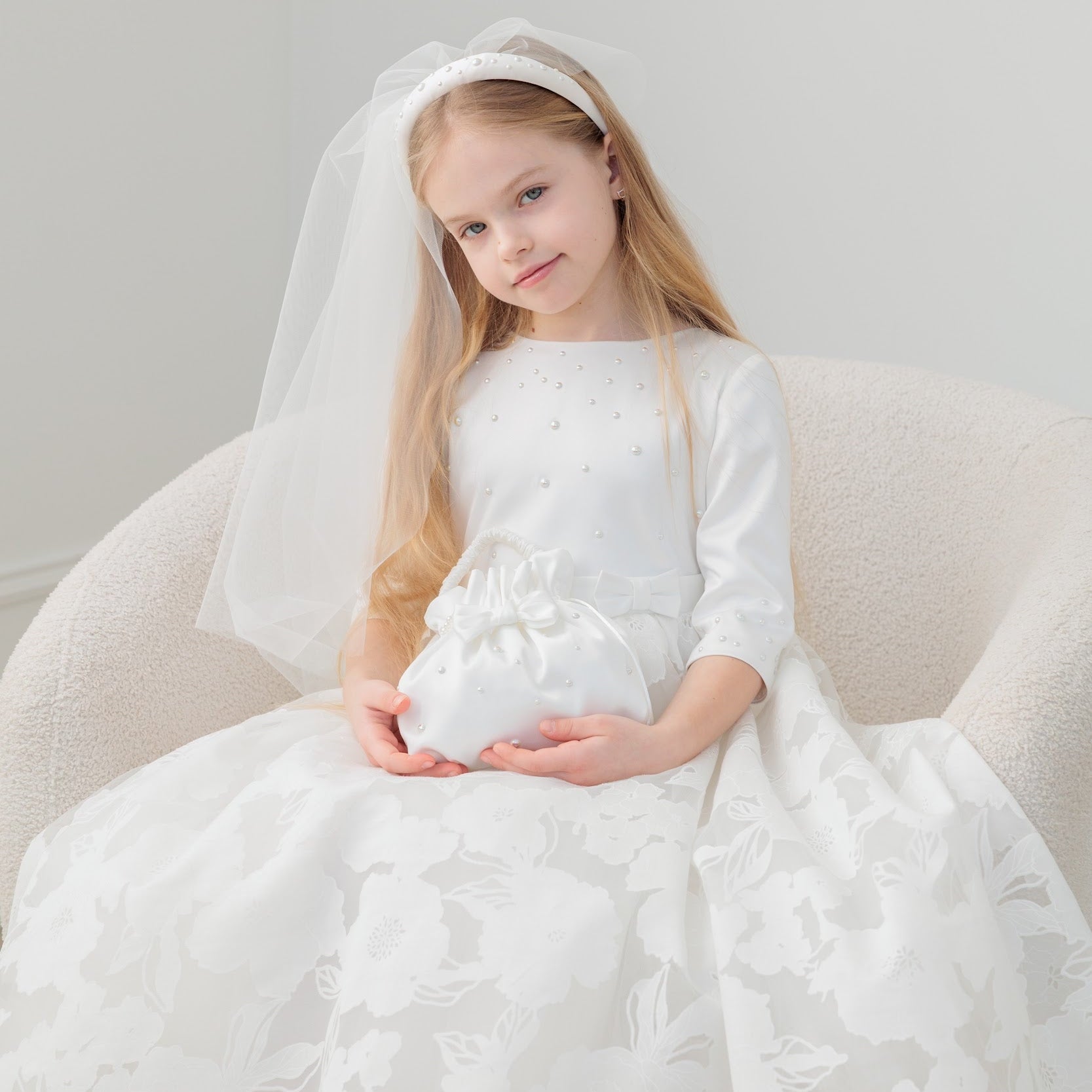 Young girl in a white communion dress with a veil sitting on a white chair.