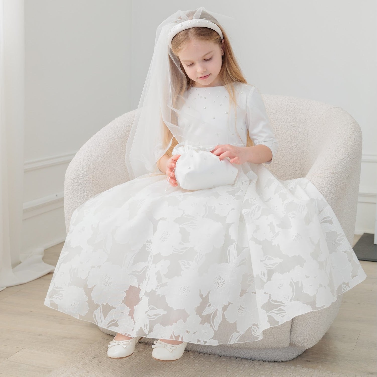 Young girl in a white lace communion dress sitting on a chair holding a white teddy bear in a bright room.