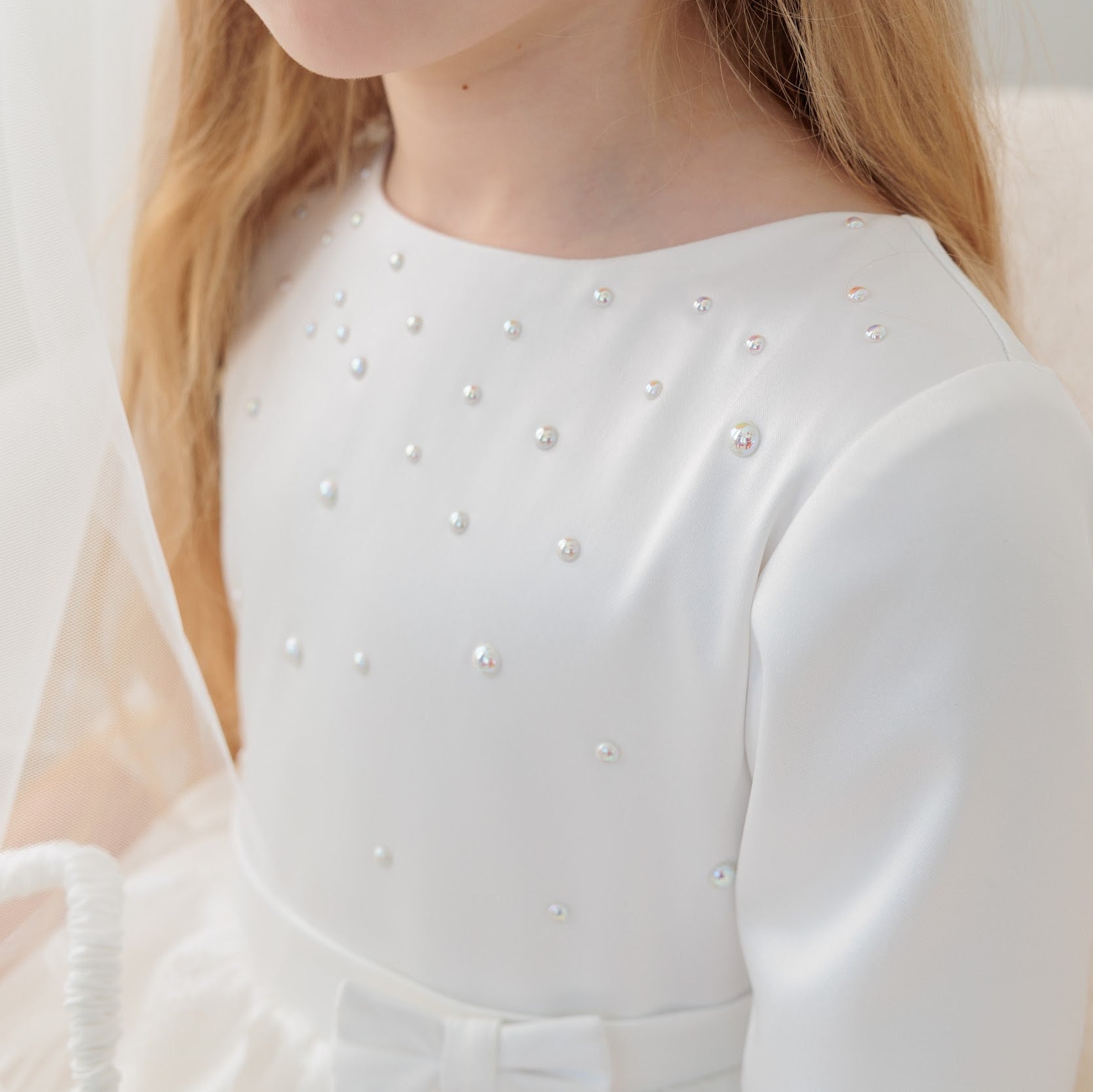 White communion dress with pearl details worn by a person, sitting on a white couch.