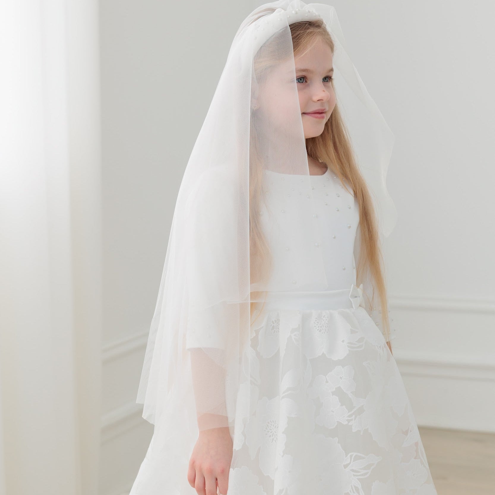 Young girl in a white communion dress with a veil against a plain background