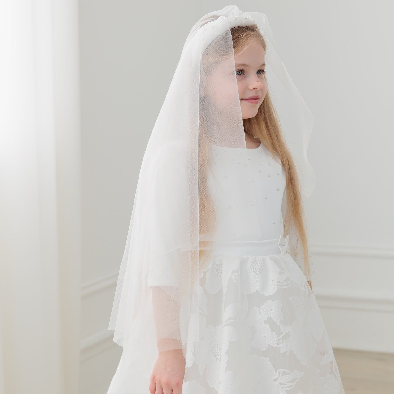 Young girl in a white dress with a veil against a plain background