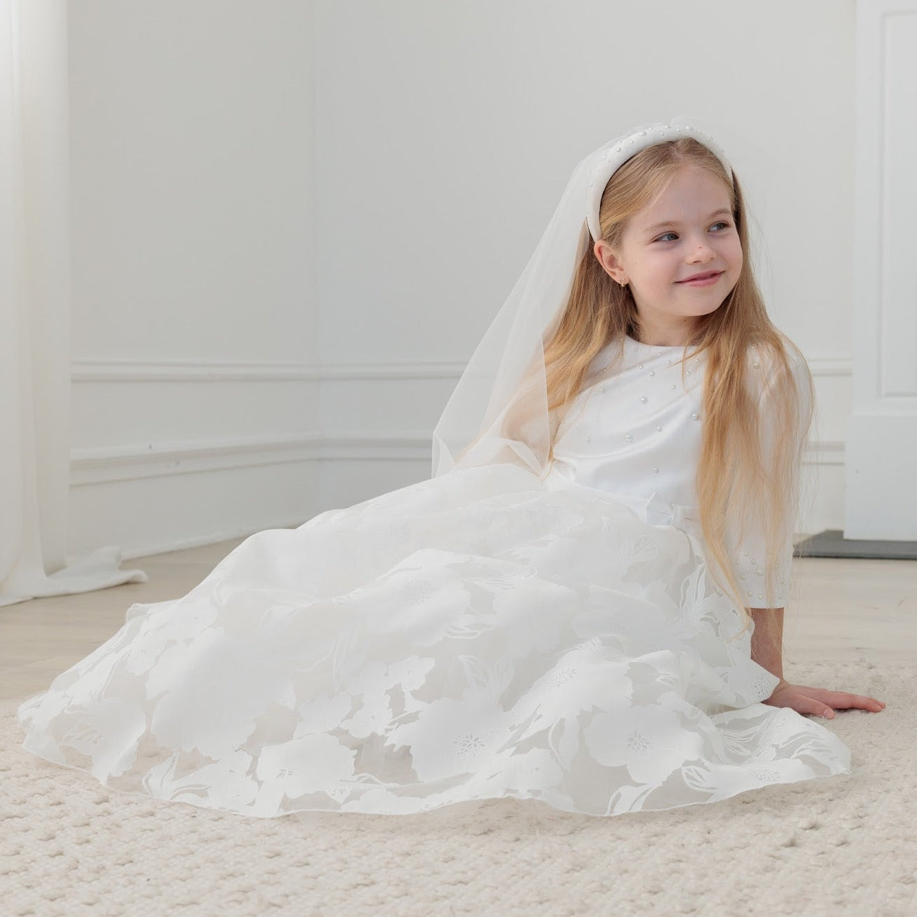 Young girl in a white communion dress sitting on a carpeted floor in a room with a fireplace.