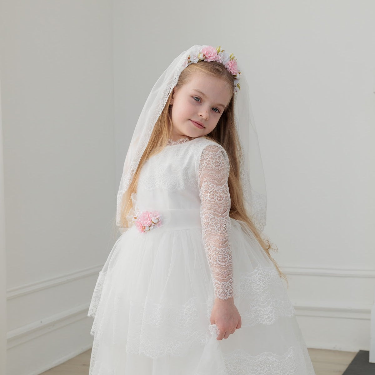 Young girl in a white dress with long sleeves and a communion veil standing in a room with white walls and a fireplace.