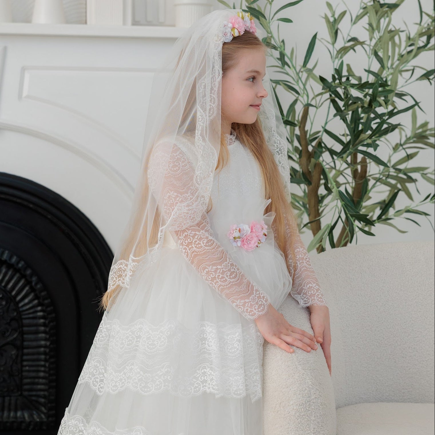 Young girl in an ivory lace communion dress with a veil standing in a room with a fireplace and plants.