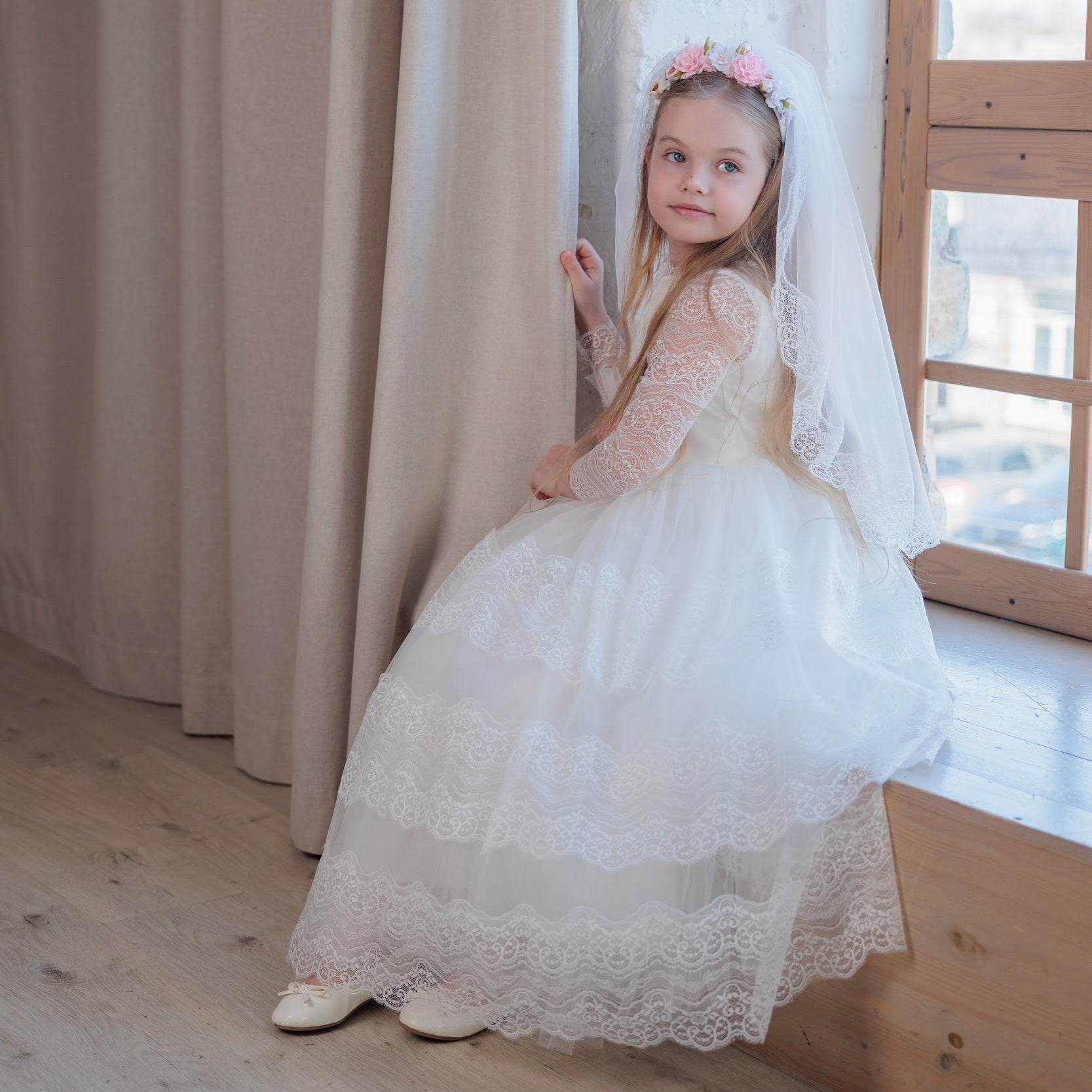 Young girl in an ivory lace communion dress standing in a room with wooden floor and large window.
