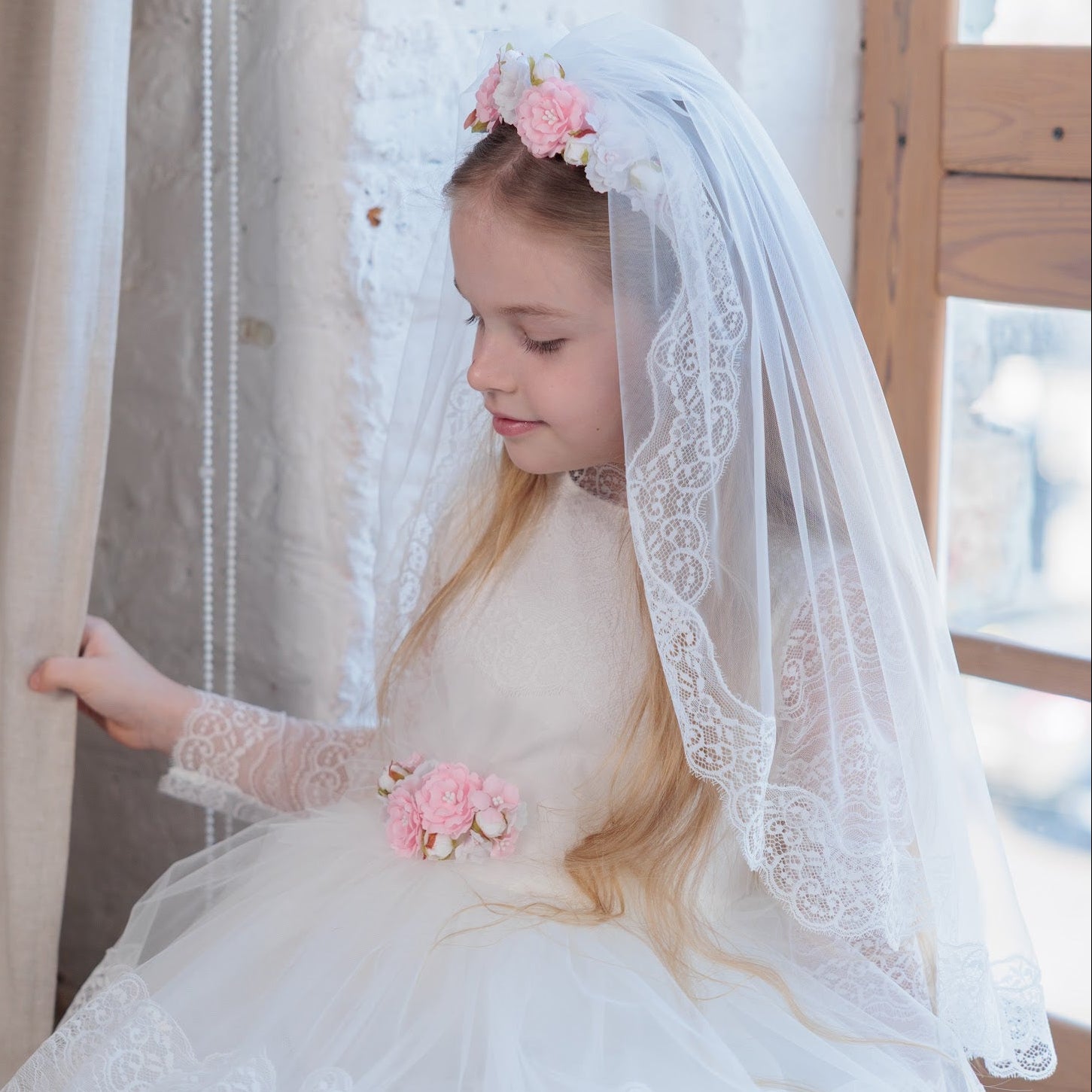 Young girl in a white dress with a communion veil and pink flowers, sitting near curtains.