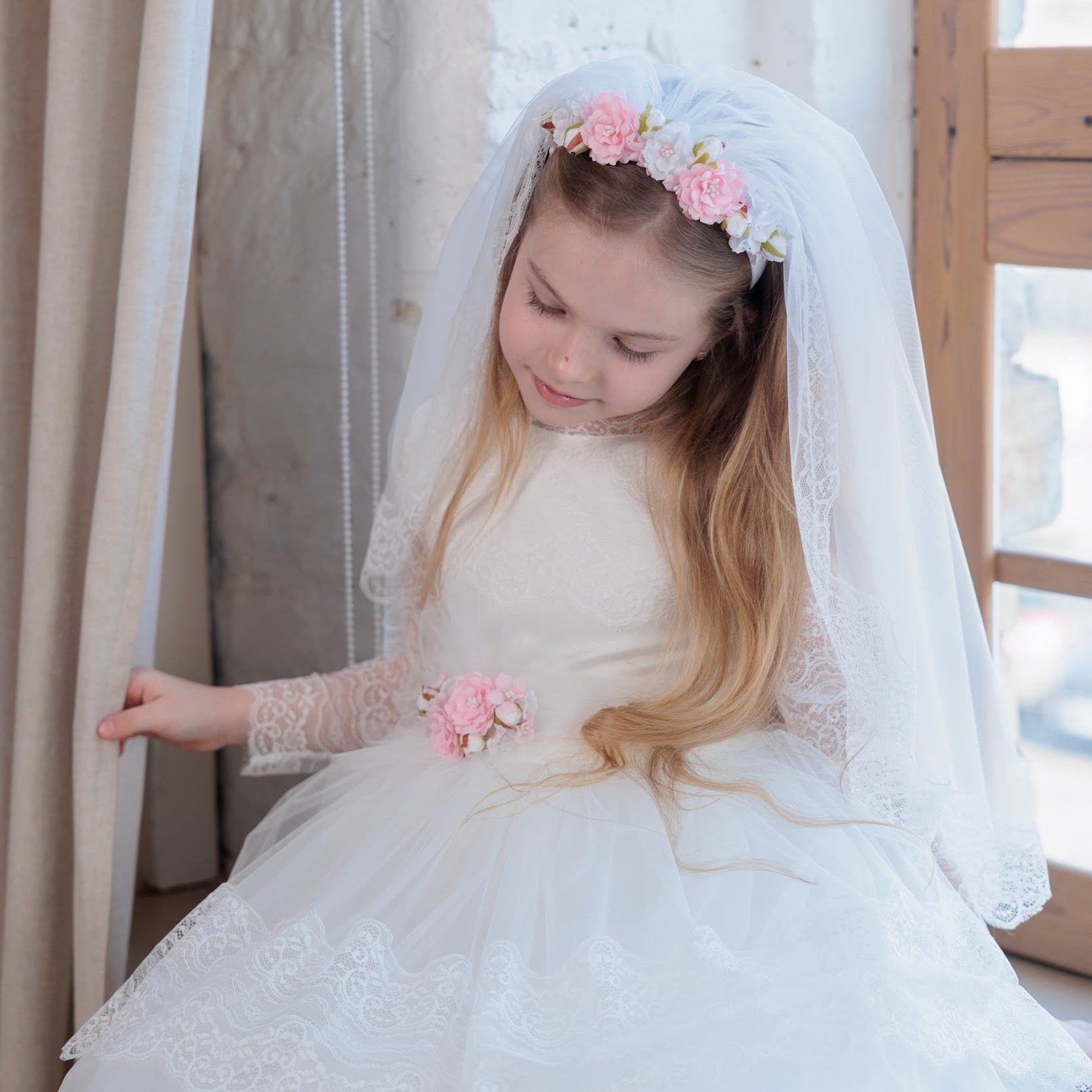 Young girl in an ivory lace communion dress with a veil and floral headband, standing indoors.