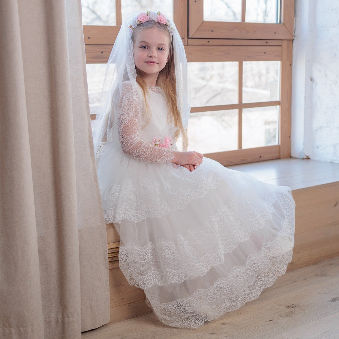 Young girl in an ivory lace communion dress with a veil sitting by a window in a bright room.