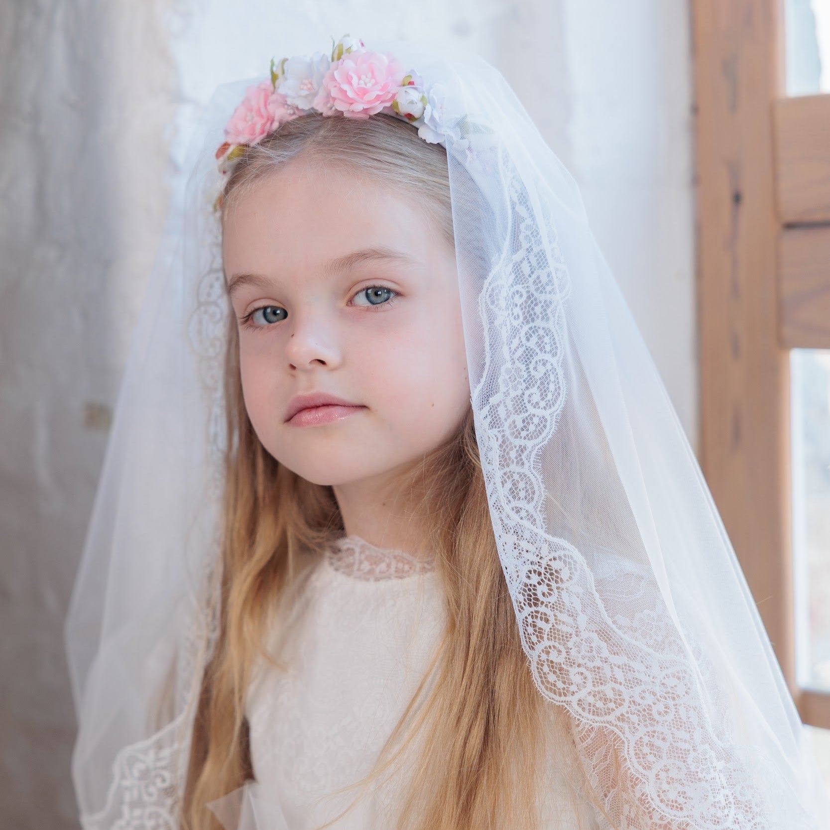 Young girl in a white dress with a veil and pink flowers, standing indoors.