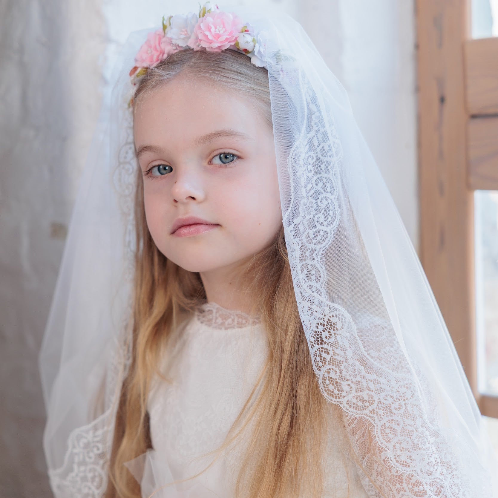 Young girl in a white dress with a communion veil and pink flowers, standing indoors.